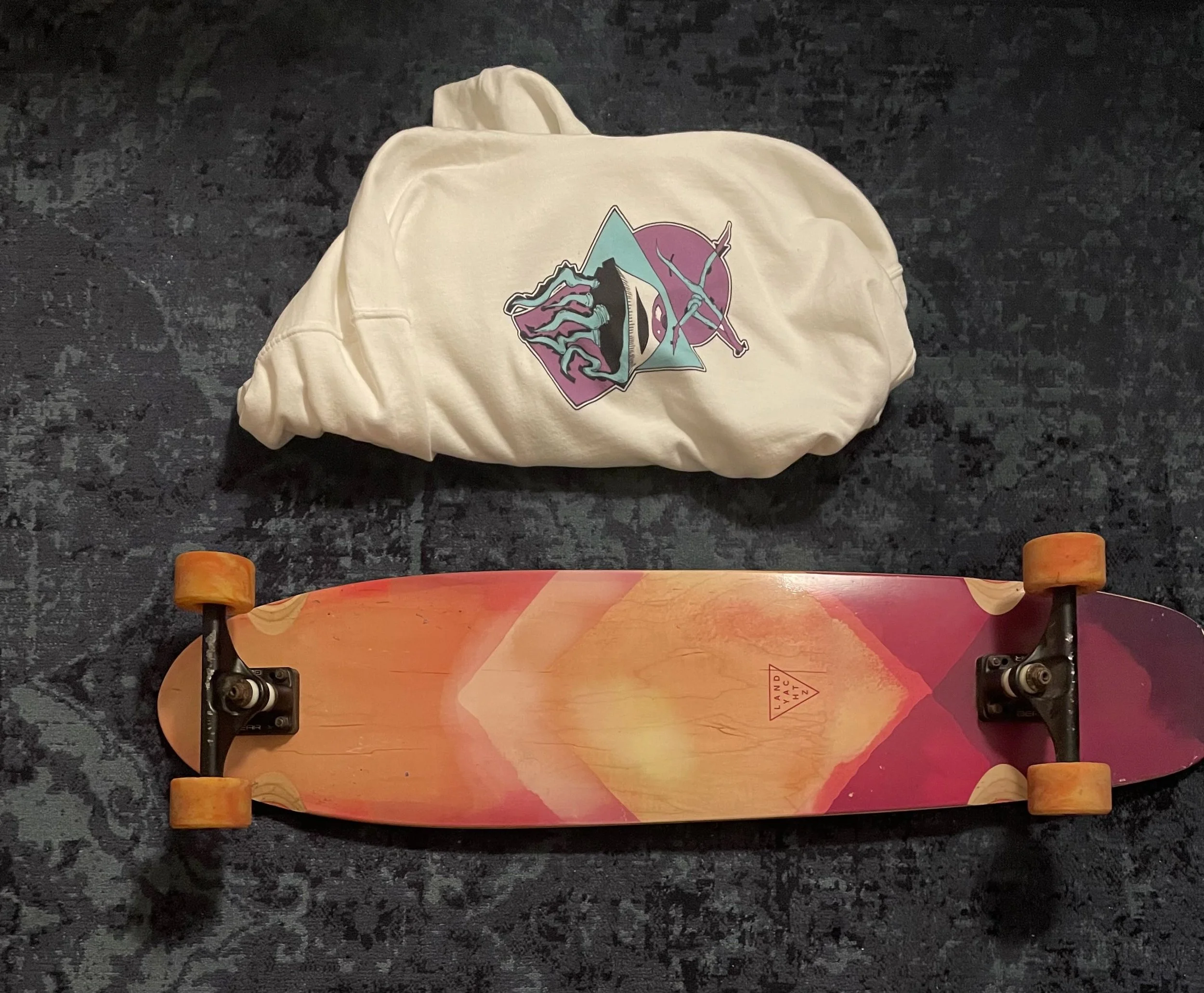 A skateboard with a colorful graphic design featuring pink, orange, and purple hues, placed on a dark textured surface next to a white hoodie with a graphic print.