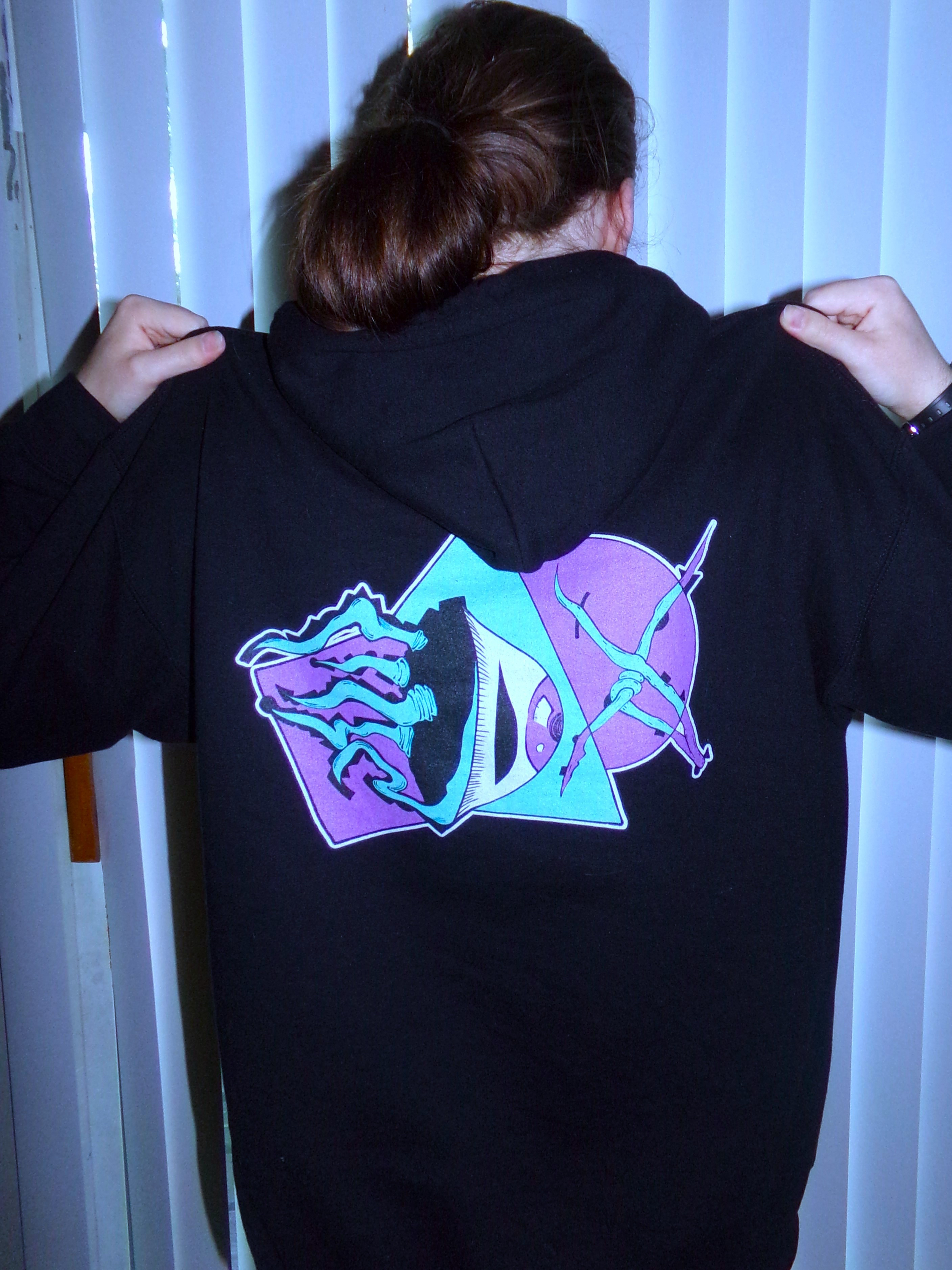 A person with brown hair, wearing a black hoodie with a neon-colored graphic on the back, holding the hoodie with both hands, standing in front of vertical white blinds.