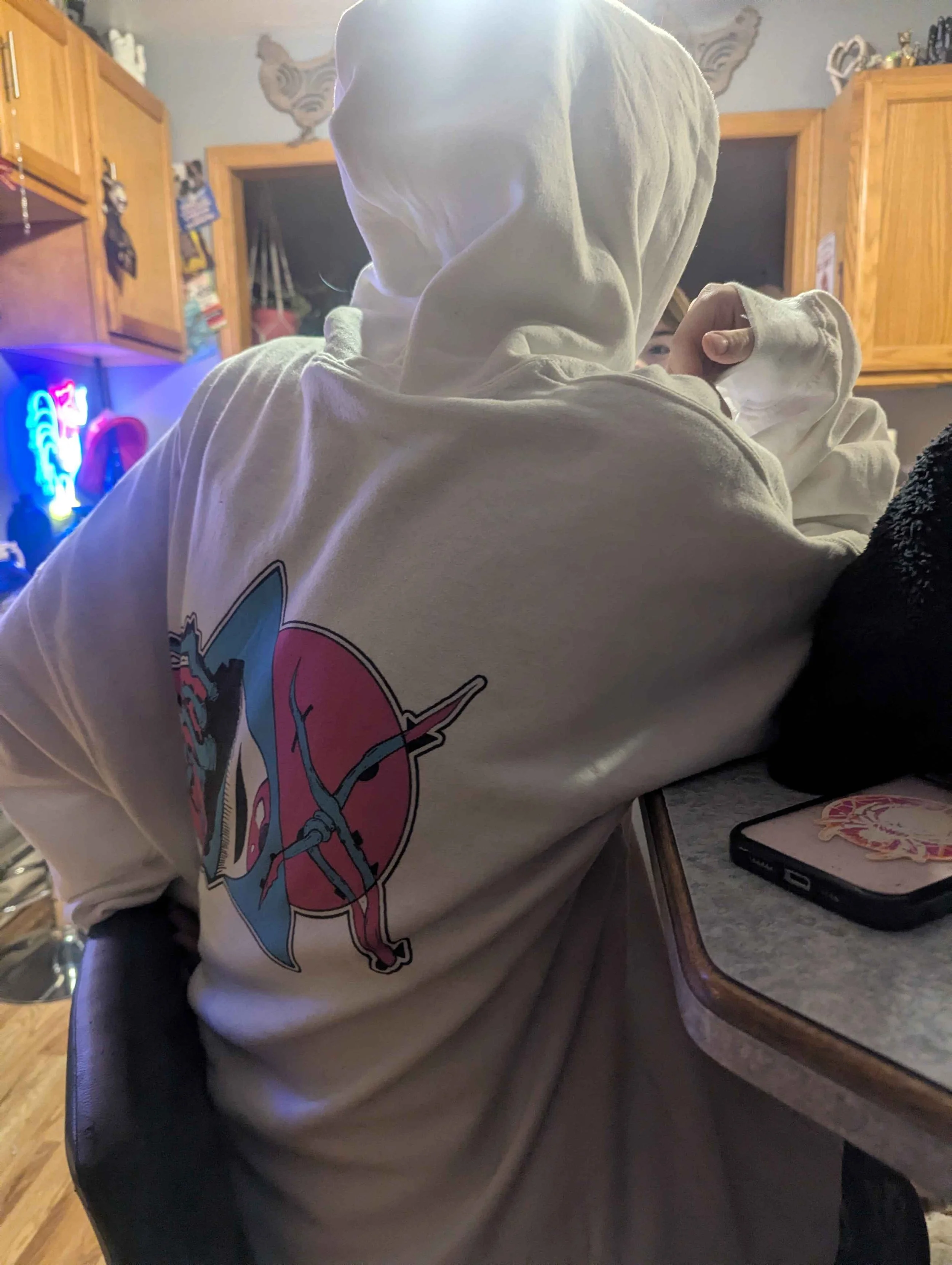 Person wearing a white hoodie with a colorful graphic design on the front, sitting in a room with wooden cabinets and a neon sign in the background.