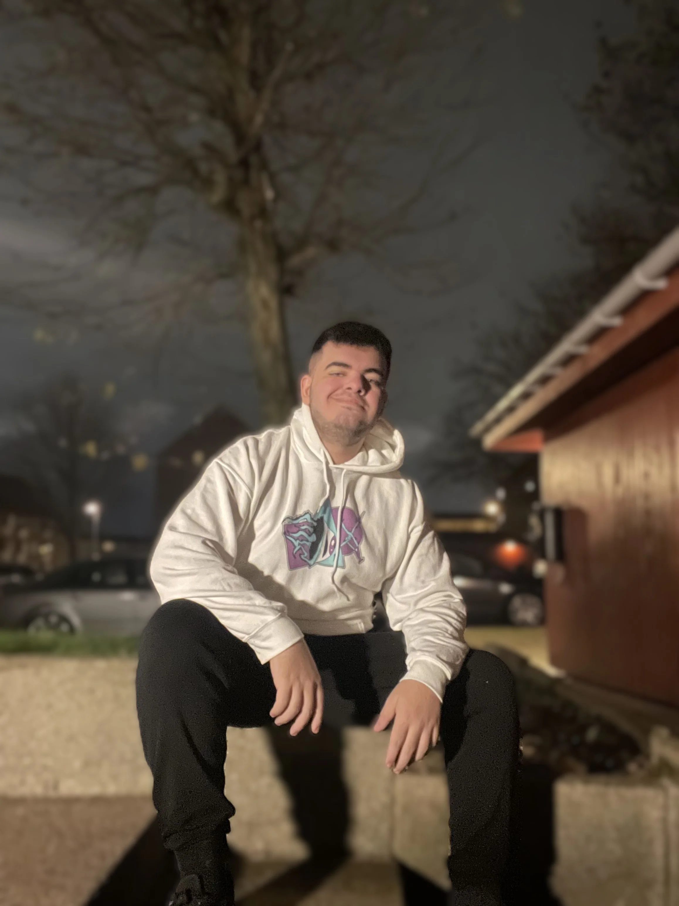 A young man with short dark hair and a scruffy beard, smiling slightly, wearing a white hoodie with a colorful graphic, sitting on a concrete surface outside at night, with bare trees and parked cars in the background.