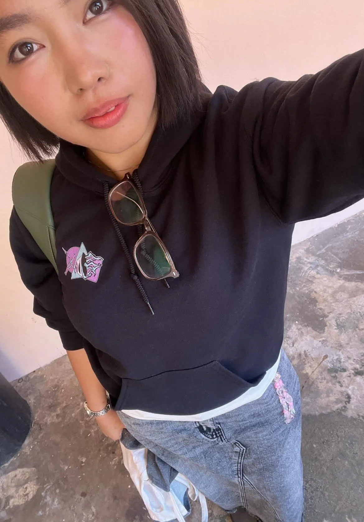 A person taking a selfie, wearing a black hoodie with a colorful logo on the chest, glasses hanging from the neckline, a green backpack strap on the shoulder, gray denim pants with embroidery, and standing on a concrete floor.