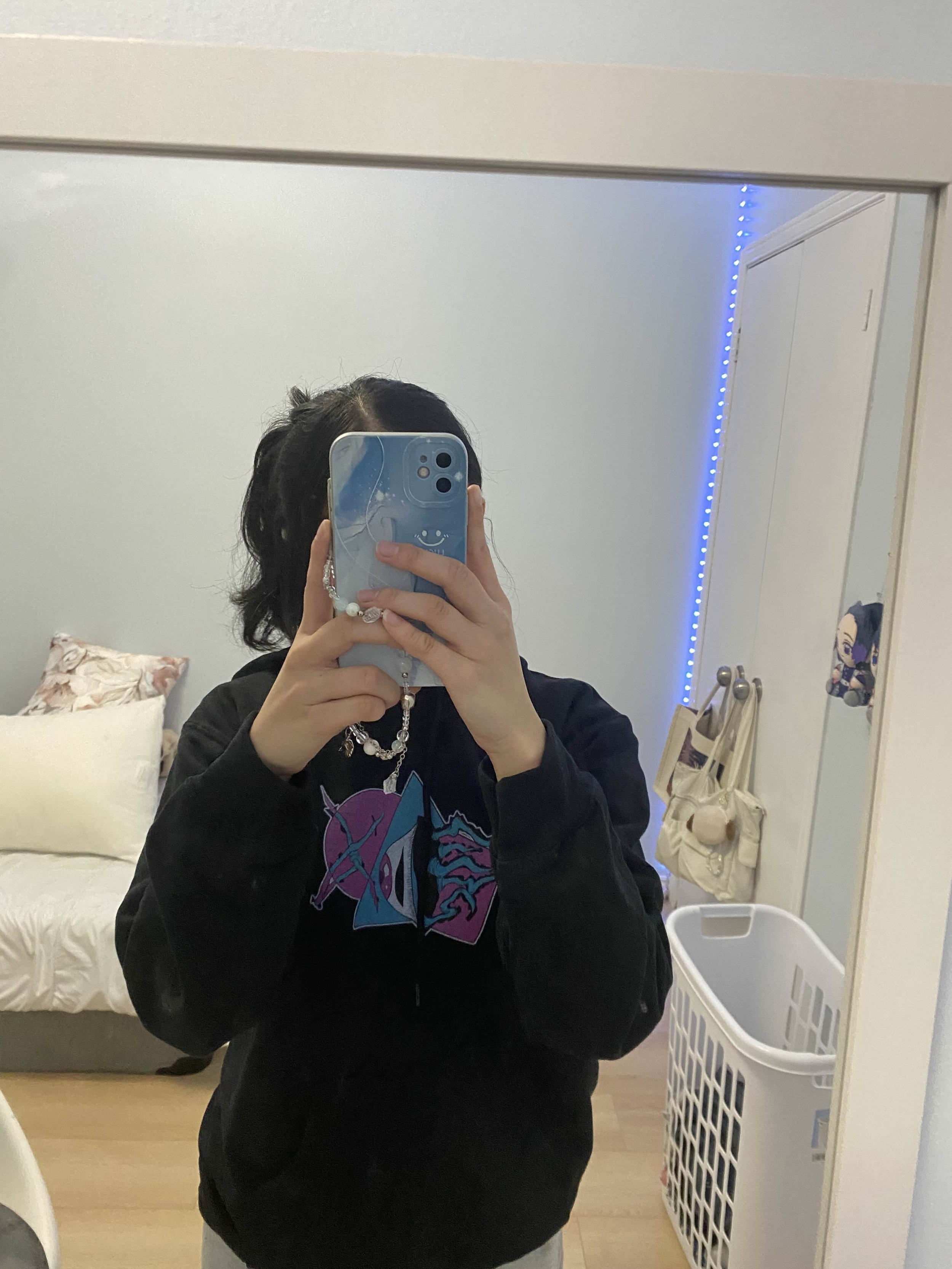 A person taking a mirror selfie in a bedroom, wearing a black hoodie with anime artwork, with a bed, pillows, a laundry basket, and a wardrobe with blue LED lights visible in the background.
