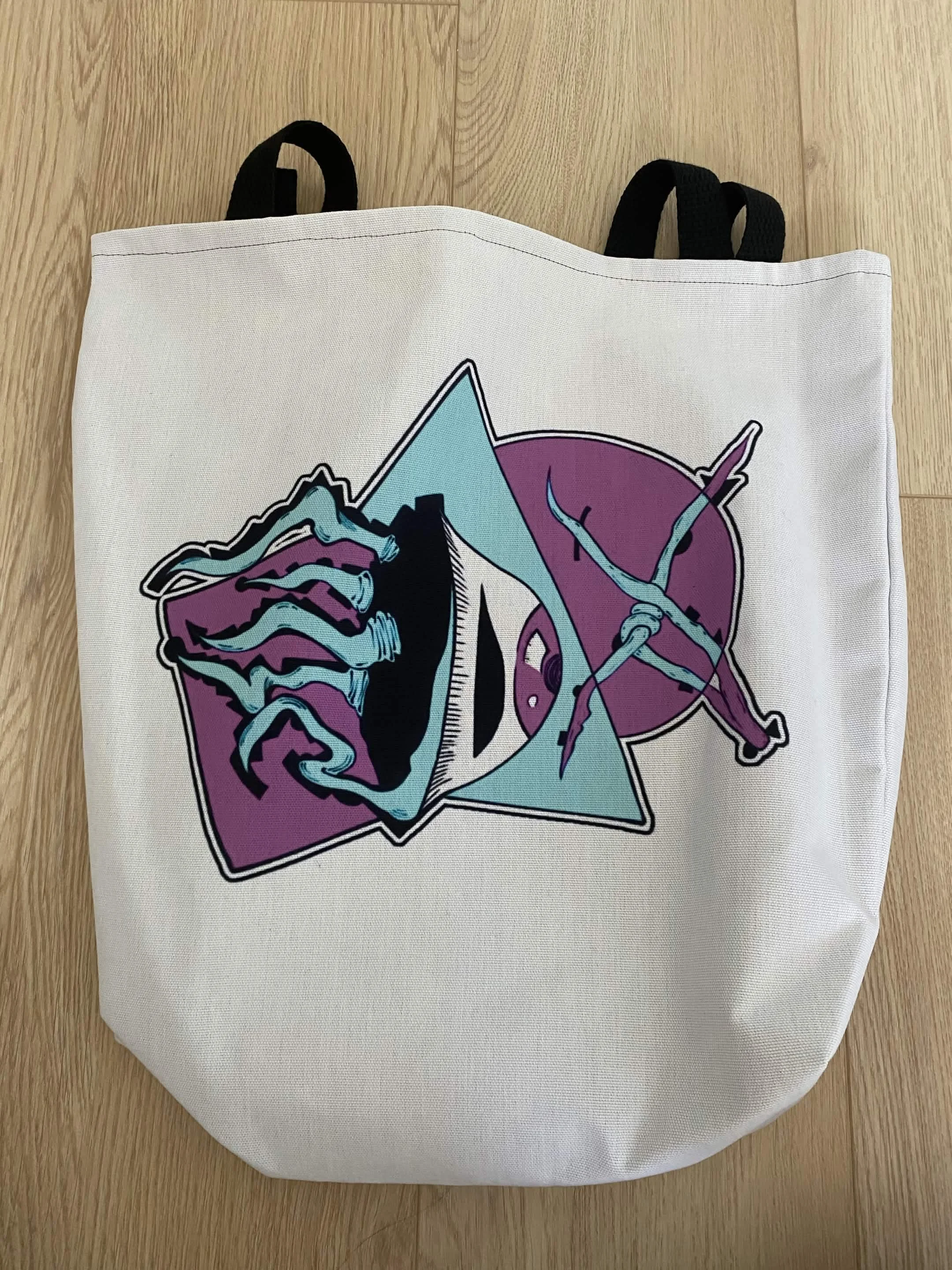 White tote bag featuring a stylized graphic design of a mysterious creature with elaborate, tentacle-like appendages, outlined with bold black lines and filled with shades of purple, blue, and teal.