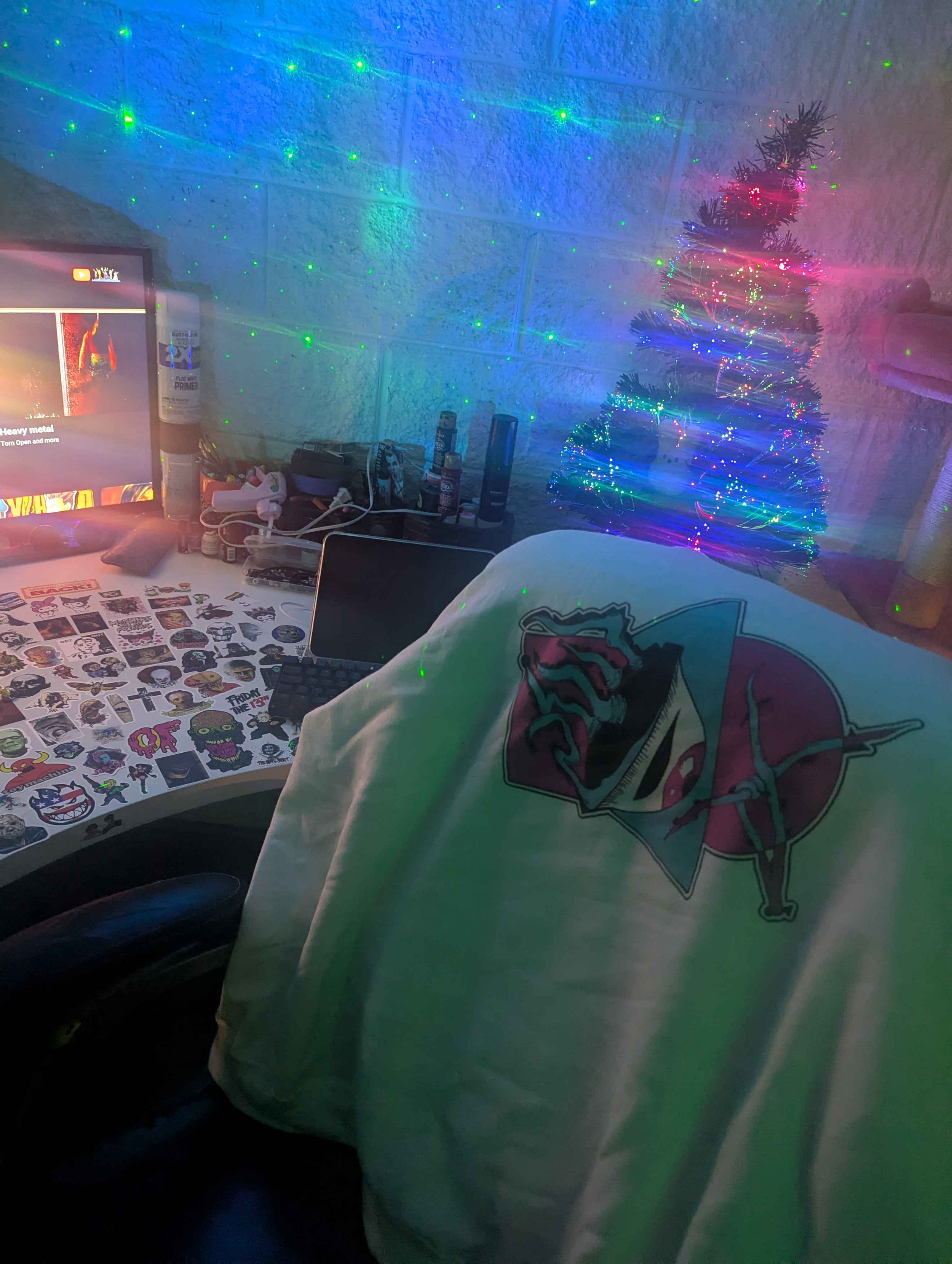 A small decorated Christmas tree with multicolored lights in a room, with a desk in the foreground showing a laptop, stickers, and various items. Green laser lights are projected on the wall and ceiling.