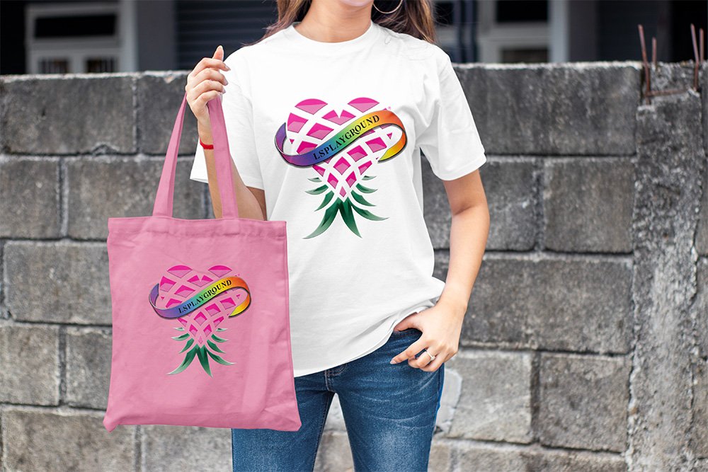 Person wearing a white t-shirt and holding a pink tote bag, both featuring a pink heart-shaped plant with a rainbow banner that reads "U.S. PLAYGROUND" against a gray stone wall.