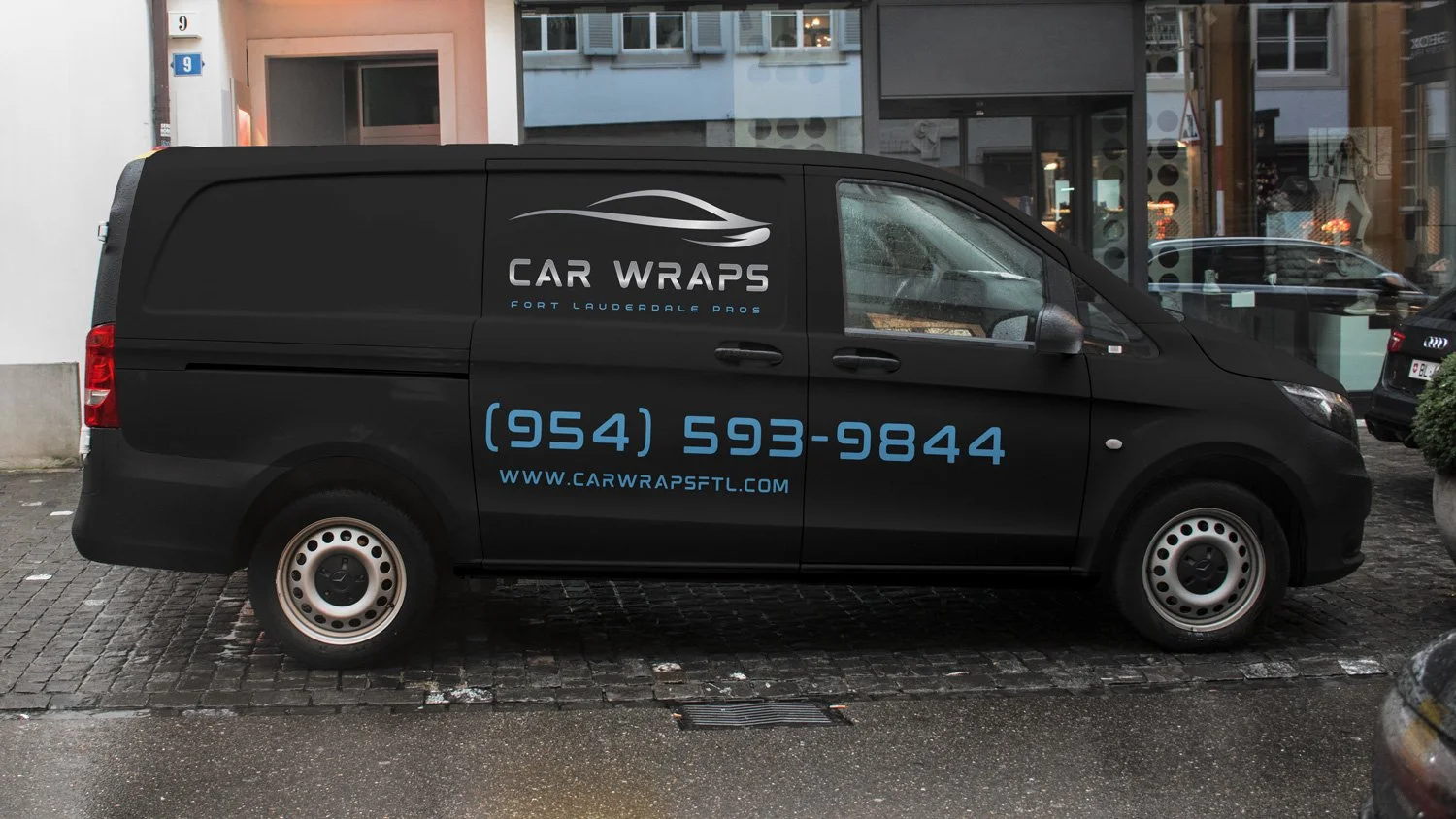Black delivery van parked on cobblestone street with advertisements for Car Wraps, displaying a logo of a stylized car silhouette and the text 'Car Wraps Fort Lauderdale Pros,' along with a phone number '(954) 593-9844' and website 'www.carwrapsftl.c