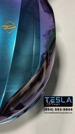 Close-up of a vehicle with Tesla wraps logo and contact information on the rear bumper, showing a shiny purple and teal color.