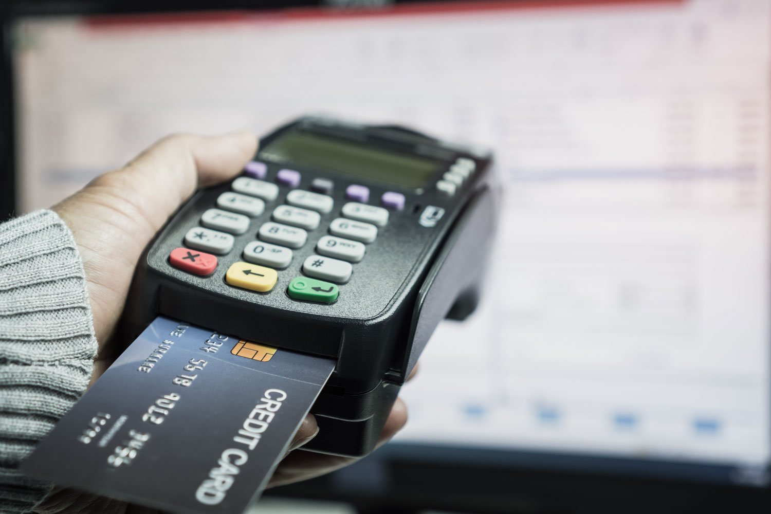 Close-up of a person's hand holding a credit card and using a point of sale terminal to make a payment.