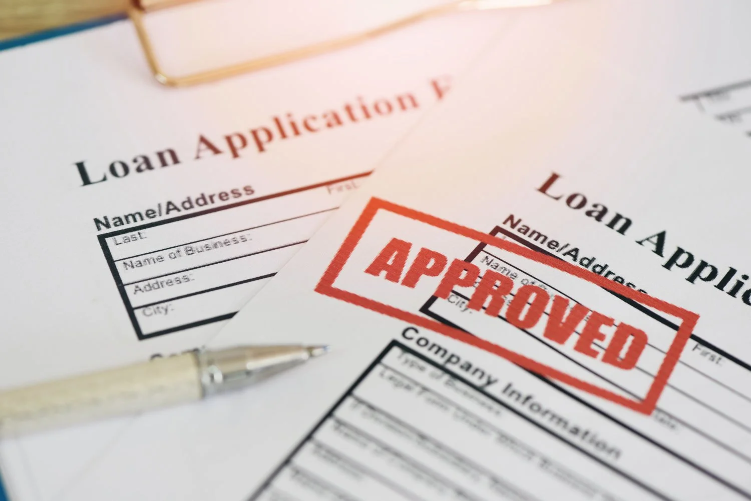 A loan application form with a red "APPROVED" stamp, partially covered by another similar form and a pen placed on top.