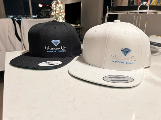 Two baseball caps, one black and one white, with embroidery logos for 'Diamond Cut Window Tinting' placed on a white countertop in a room with a TV, Christmas tree, and city lights visible in the background.