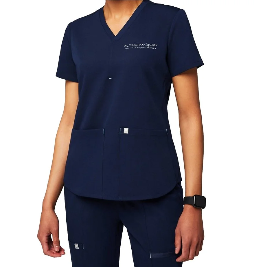 A woman wearing dark blue medical scrubs with a name tag 'Dr. Christiana Marron, Doctor of Physical Therapy' on the top and small labels on the pants. She has a black wristwatch on her left wrist.