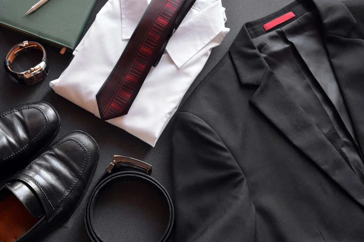 Flat lay of black dress shoes, black suit, white shirt, green notebook, wristwatch, black belt, and a measuring tape on a gray background.