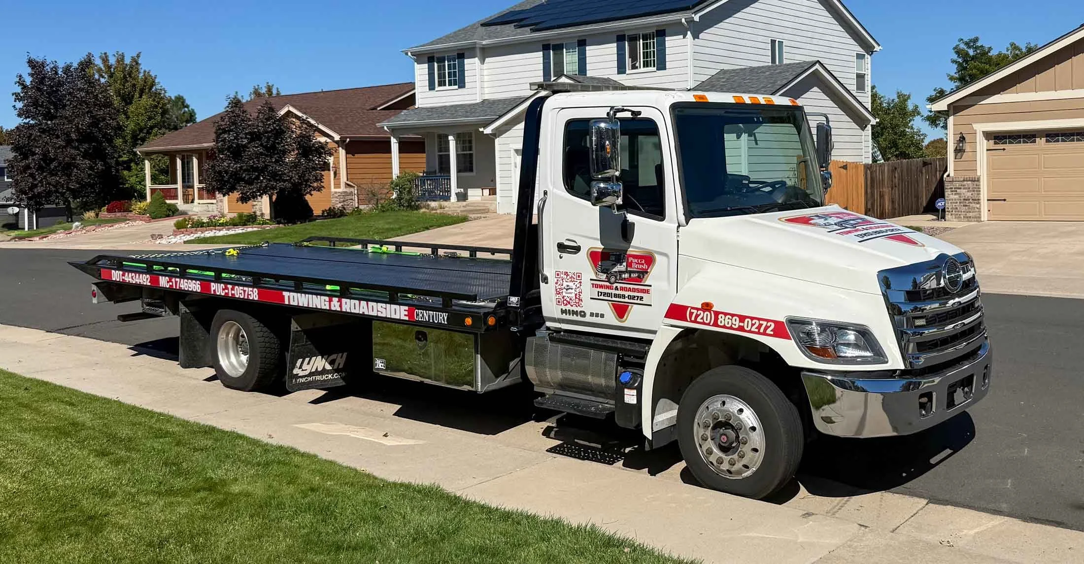 A white tow truck parked on a residential street with houses, trees, and a driveway in the background.