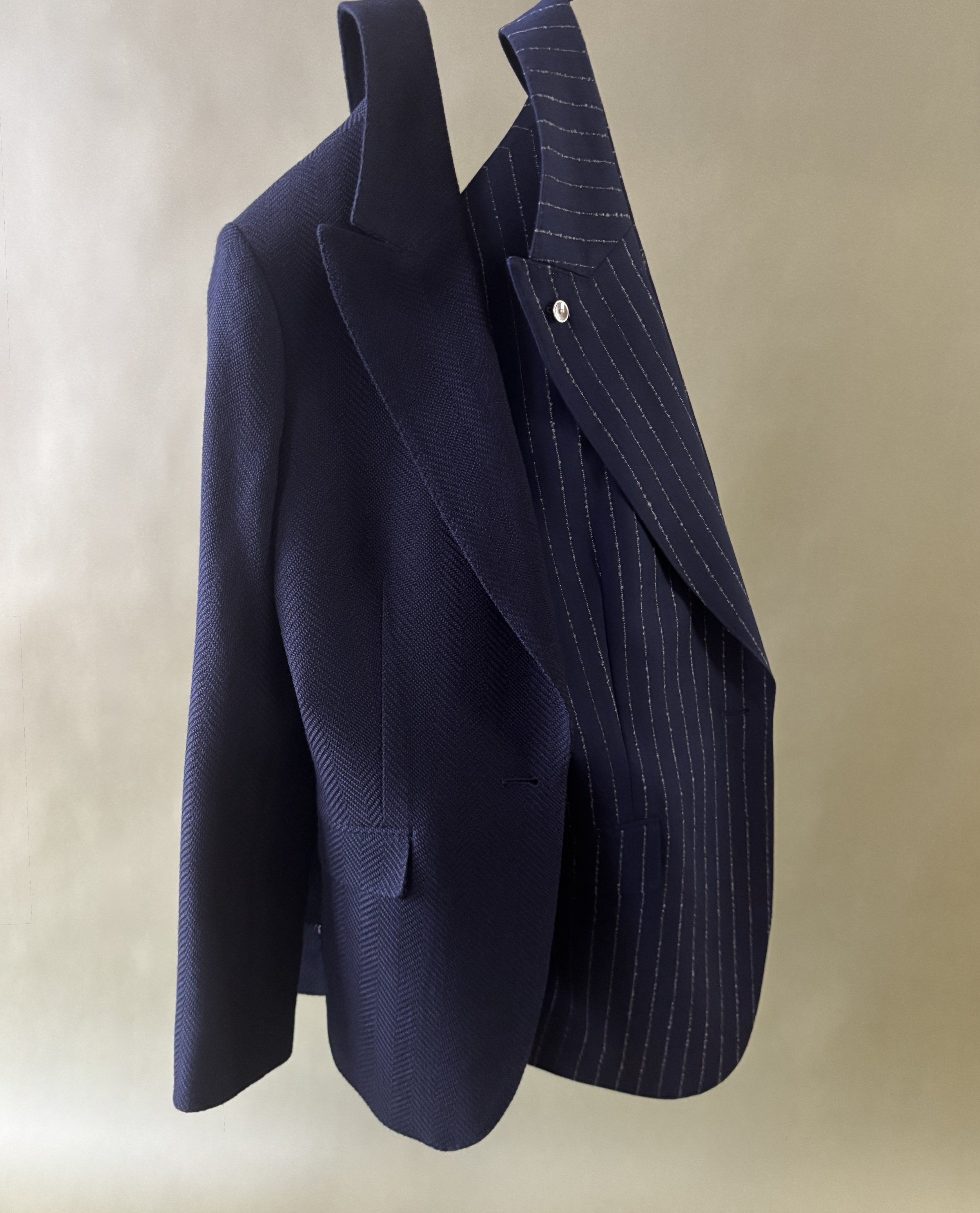 Two custom made women's blazers-one navy herringbone and one navy with white boucle stripe in a peak lapel with a mother of pearl lapel pin hanging in front of a green backdrop.