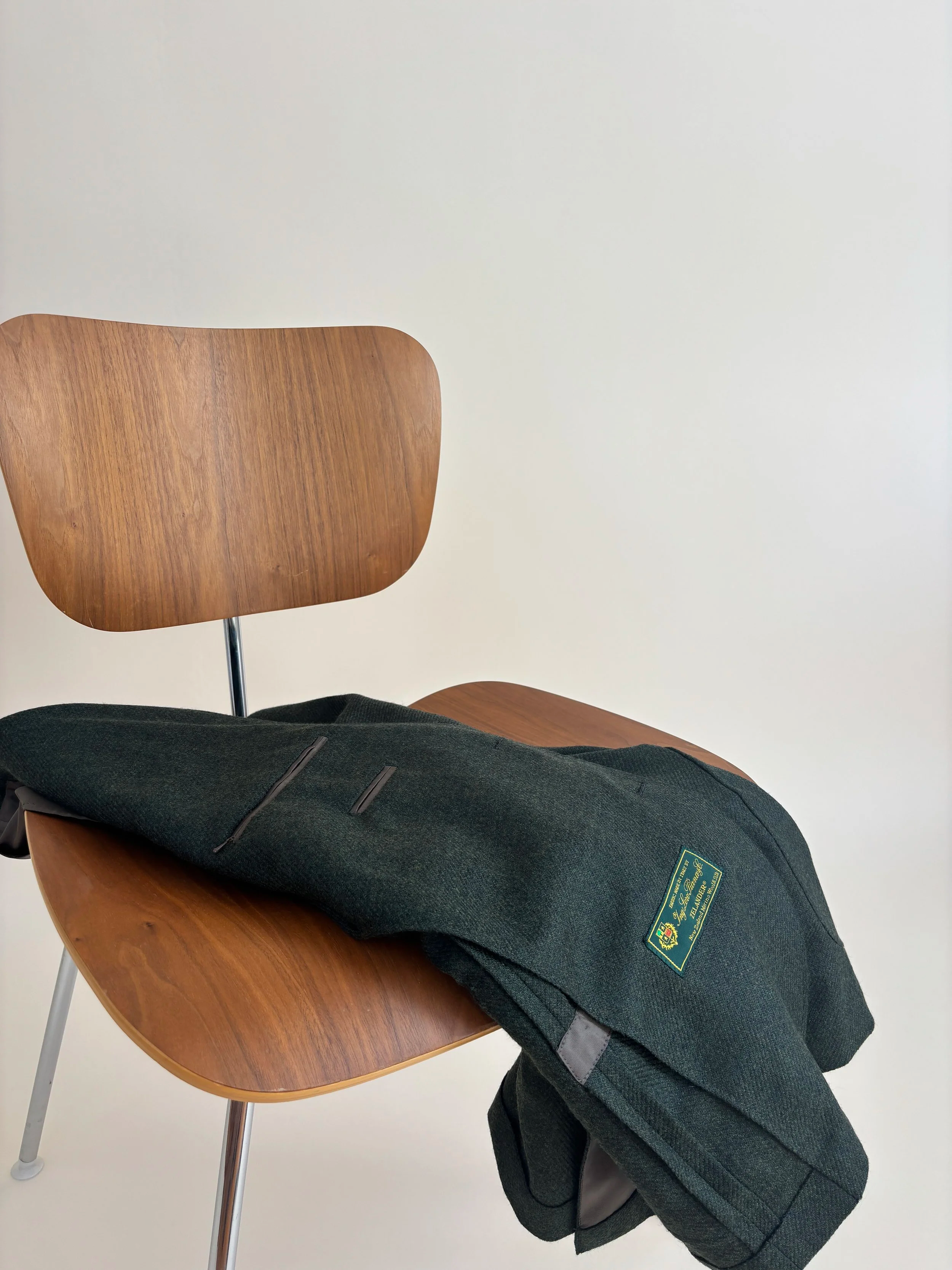 Wooden herman miller mcm chair with a hunter green flannel Loro Piana blazer draped over it, against a tan background.