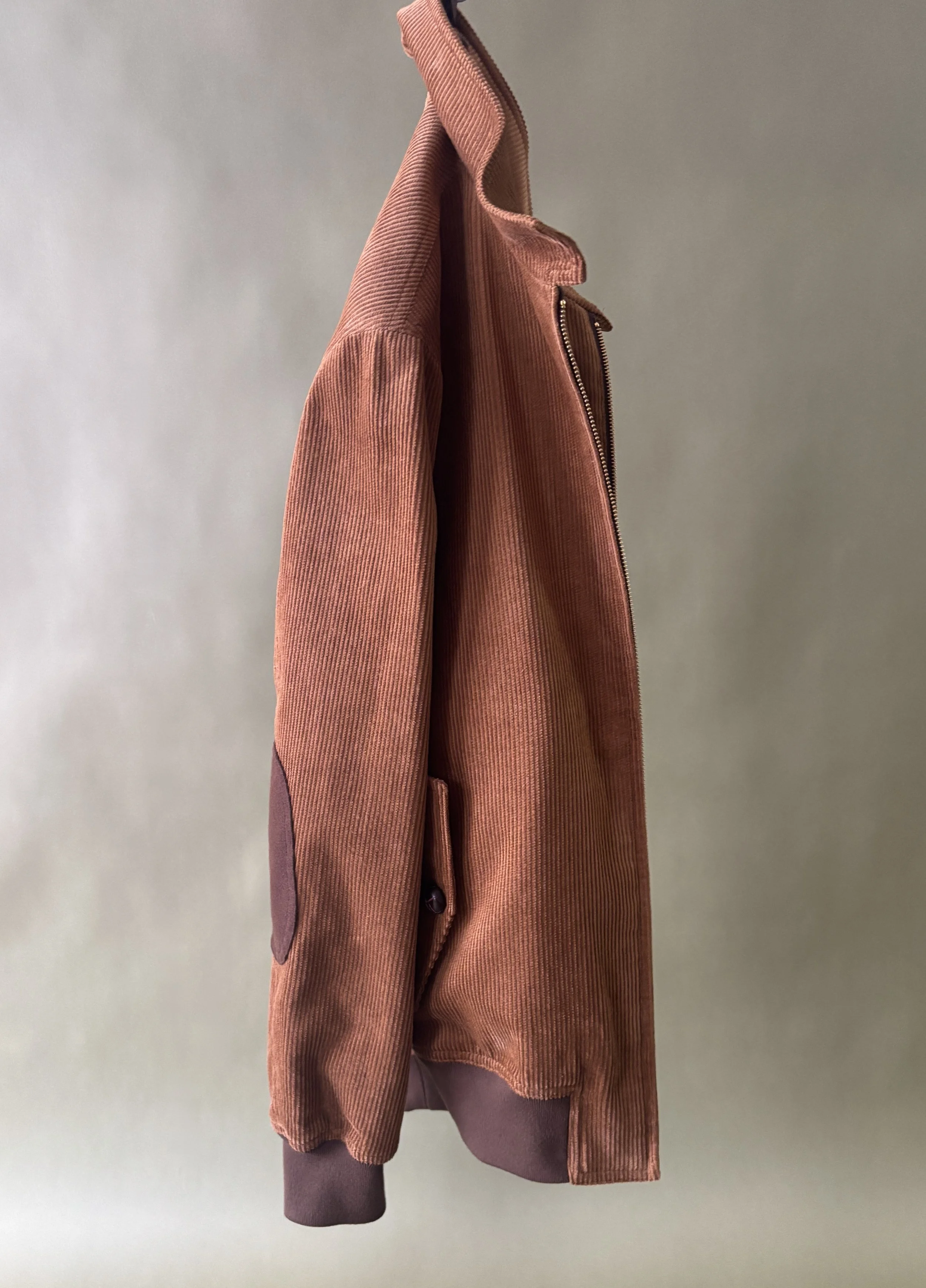 Custom made caramel brown corduroy bomber jacket with brown suede elbow patches and brown leather buttons against a green background.