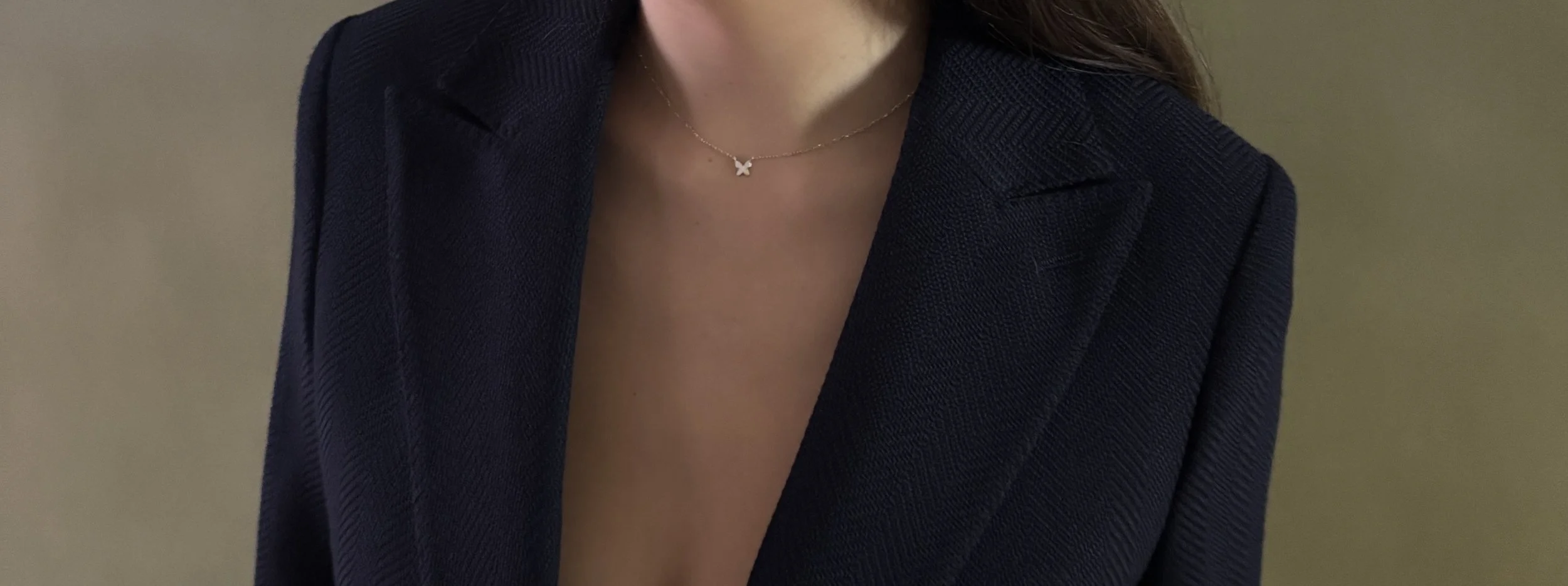 A close-up of a custom made Womens navy herringbone blazer with a necklace charm against a green background.