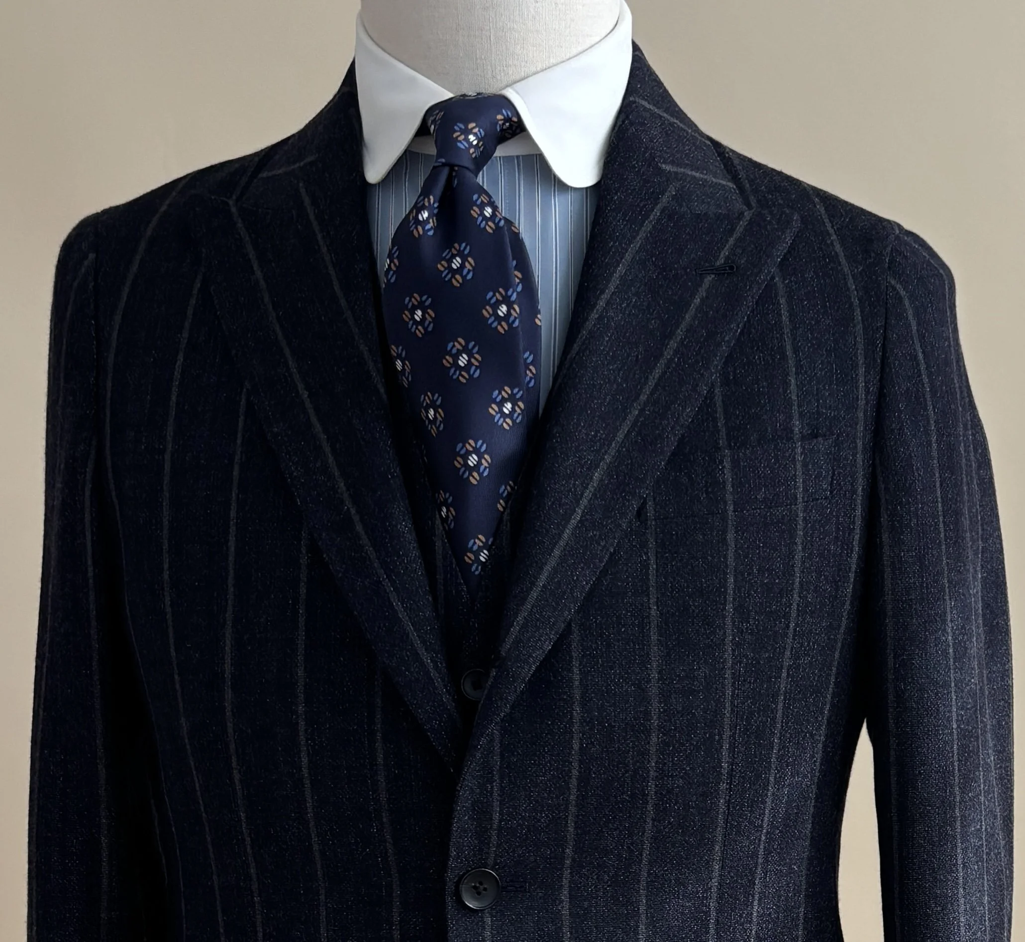 A close-up of a custom made three piece navy chalk stripe suit with a contrast club collar shirt and blue Geoff Nicholson silk necktie with medallions on a mannequin.