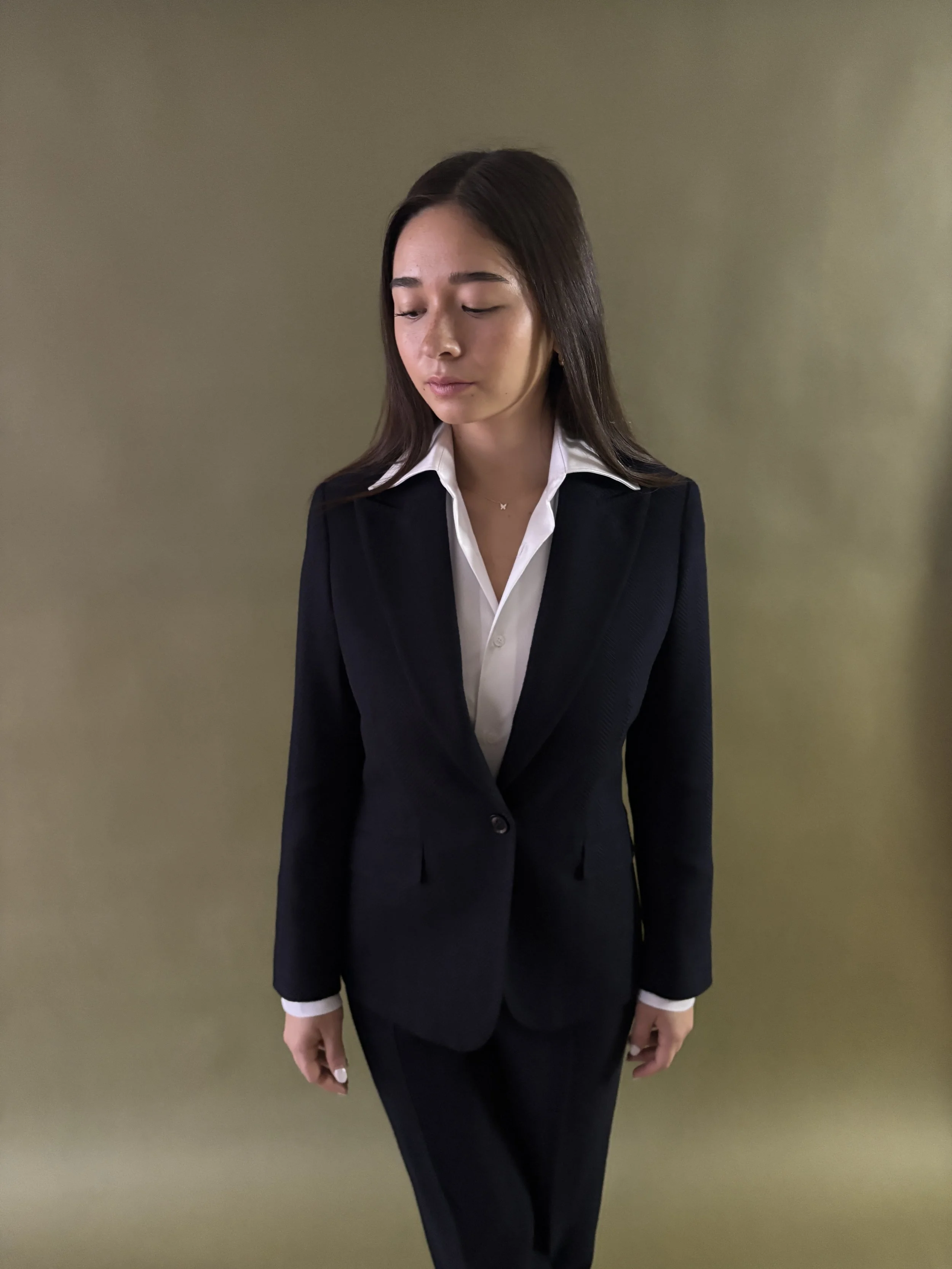 A custom made women's suit in navy herringbone knit fabric by Stylbiella, with custom made women's white dress shirt against a green background.