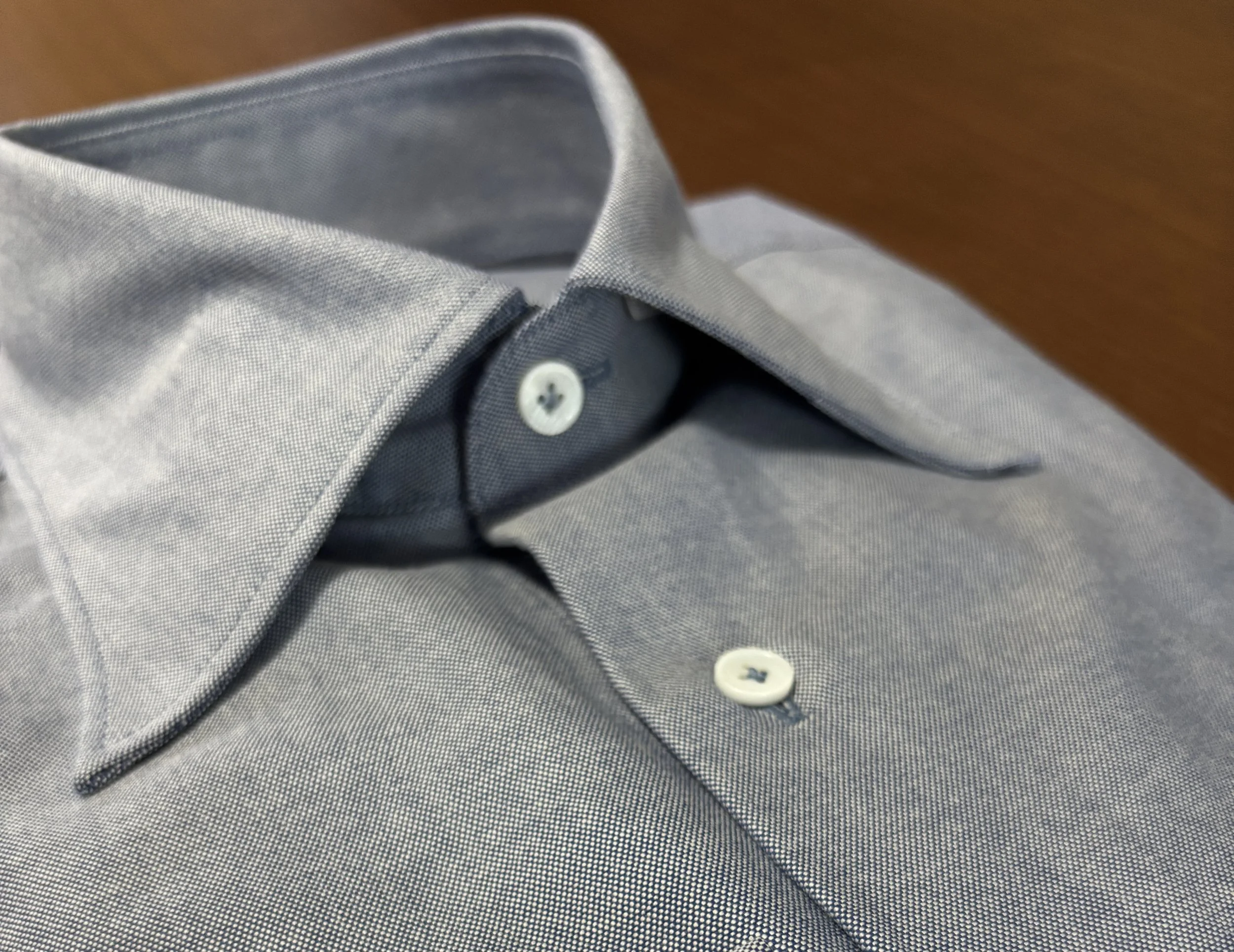 A close up of a custom made indigo blue chambray dress shirt with shell/mother of pearl buttons on walnut wood table.