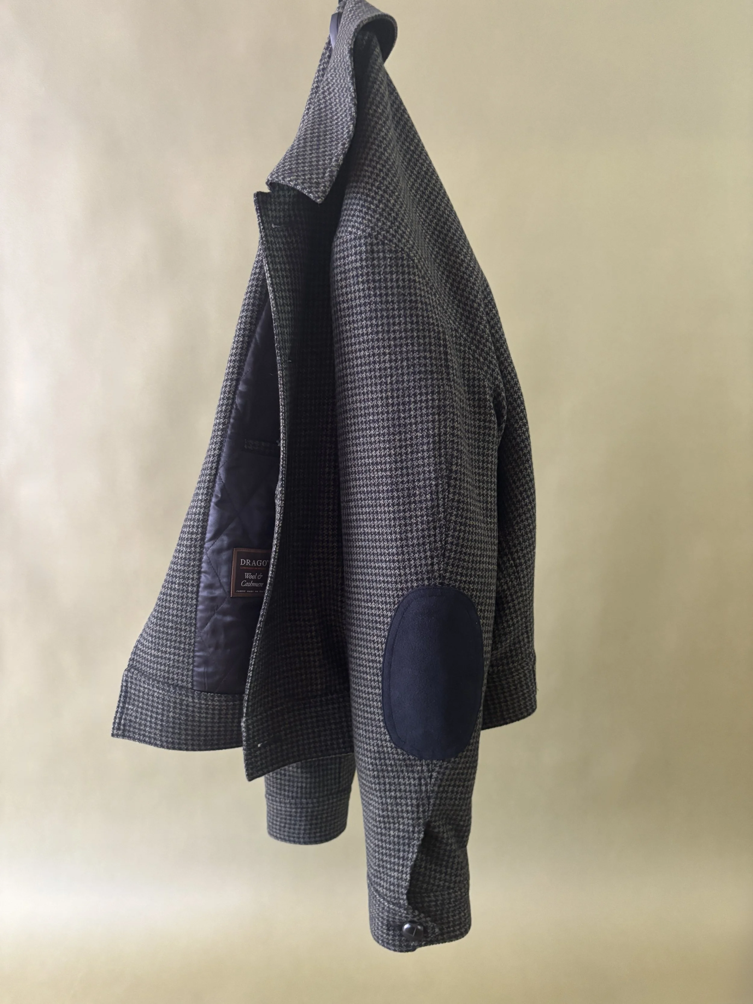 A custom made green and black houndstooth Drago wool & cashmere bomber jacket with black suede elbow patches hanging against a green backdrop.