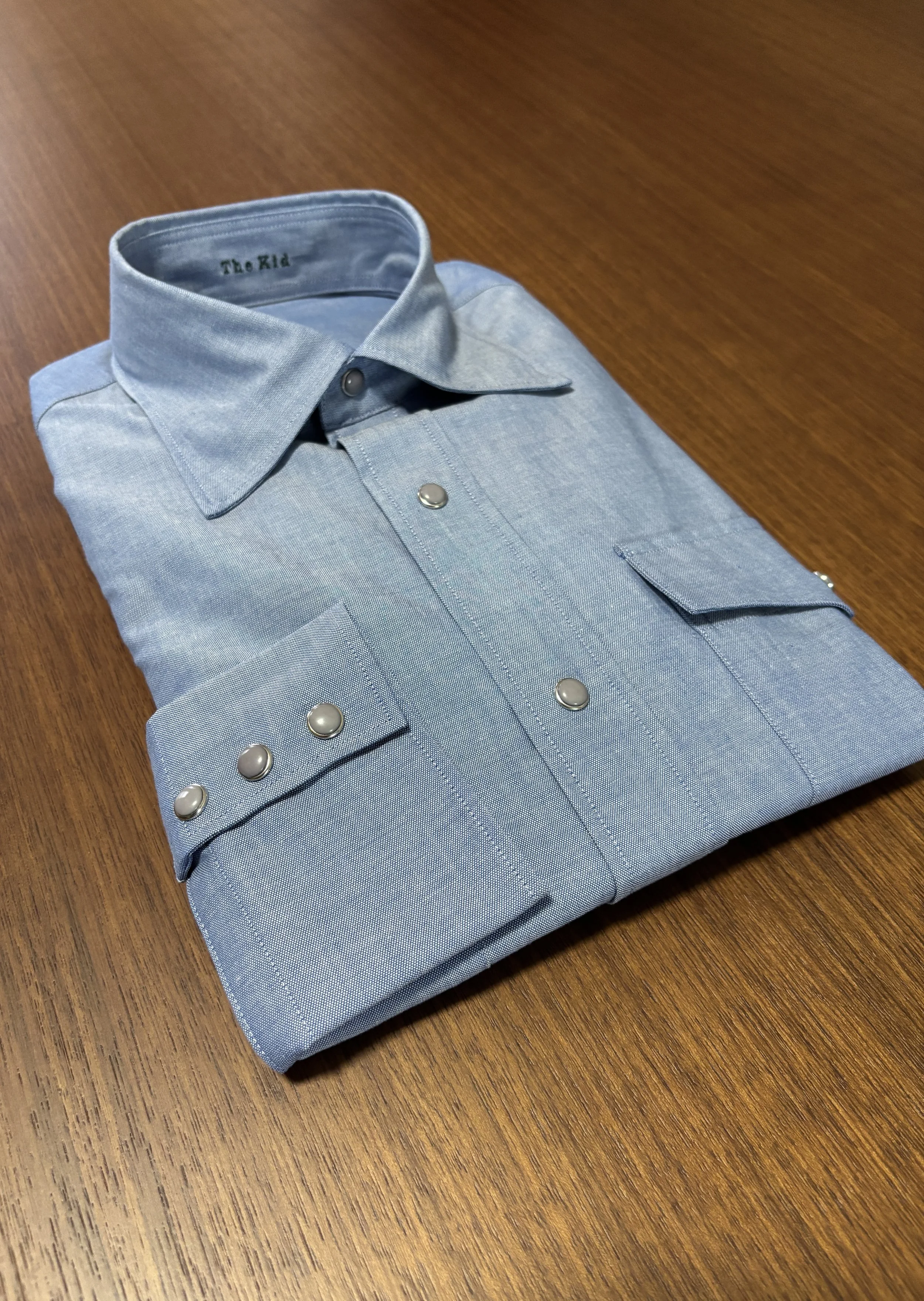 A custom made blue chambray western shirt with snaps on a walnut wooden table.