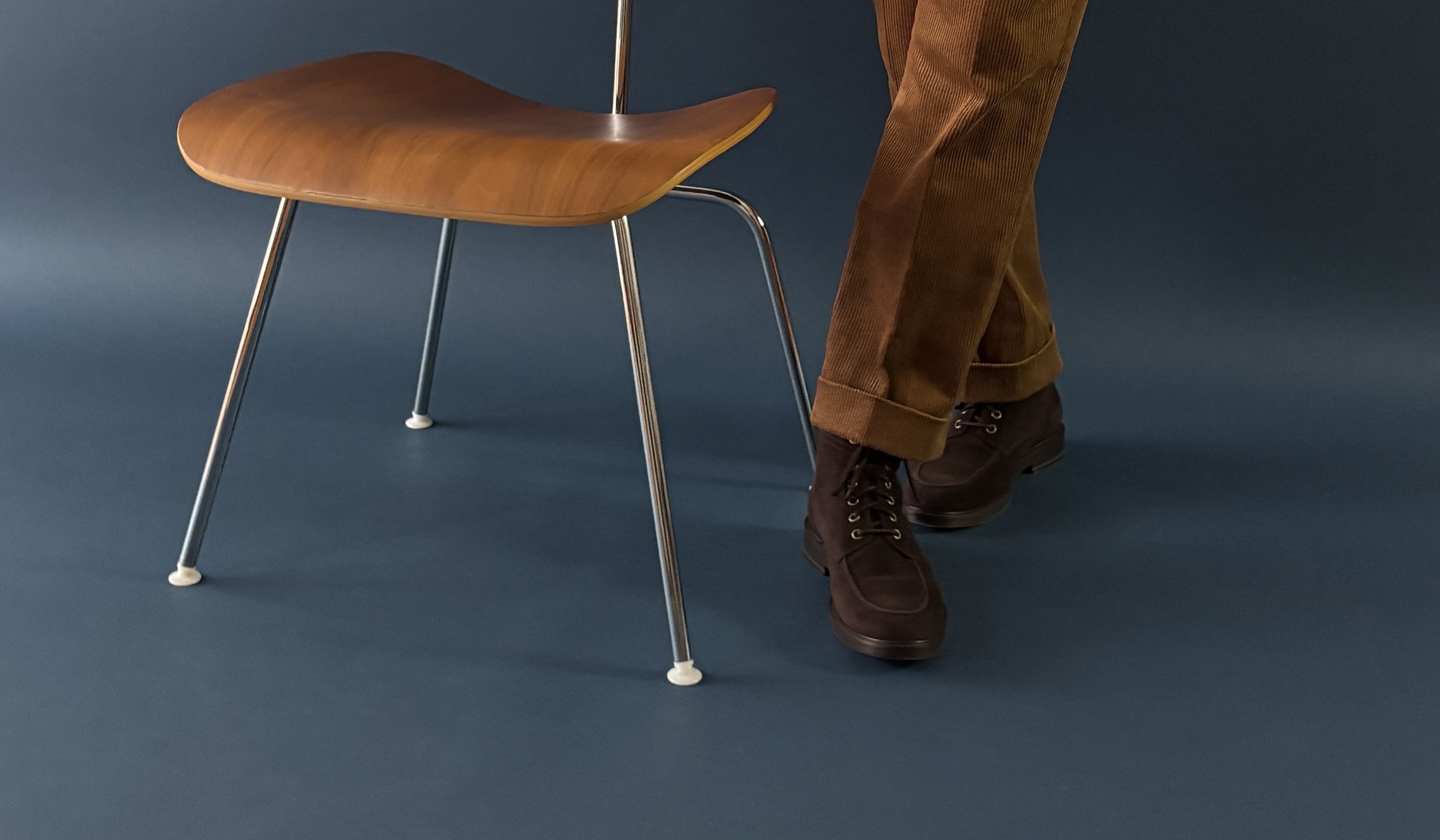 A close-up of custom made caramel brown corduroy trousers with a cuff and brown suede boots with wooden Herman Miller chair and an indigo blue backdrop.