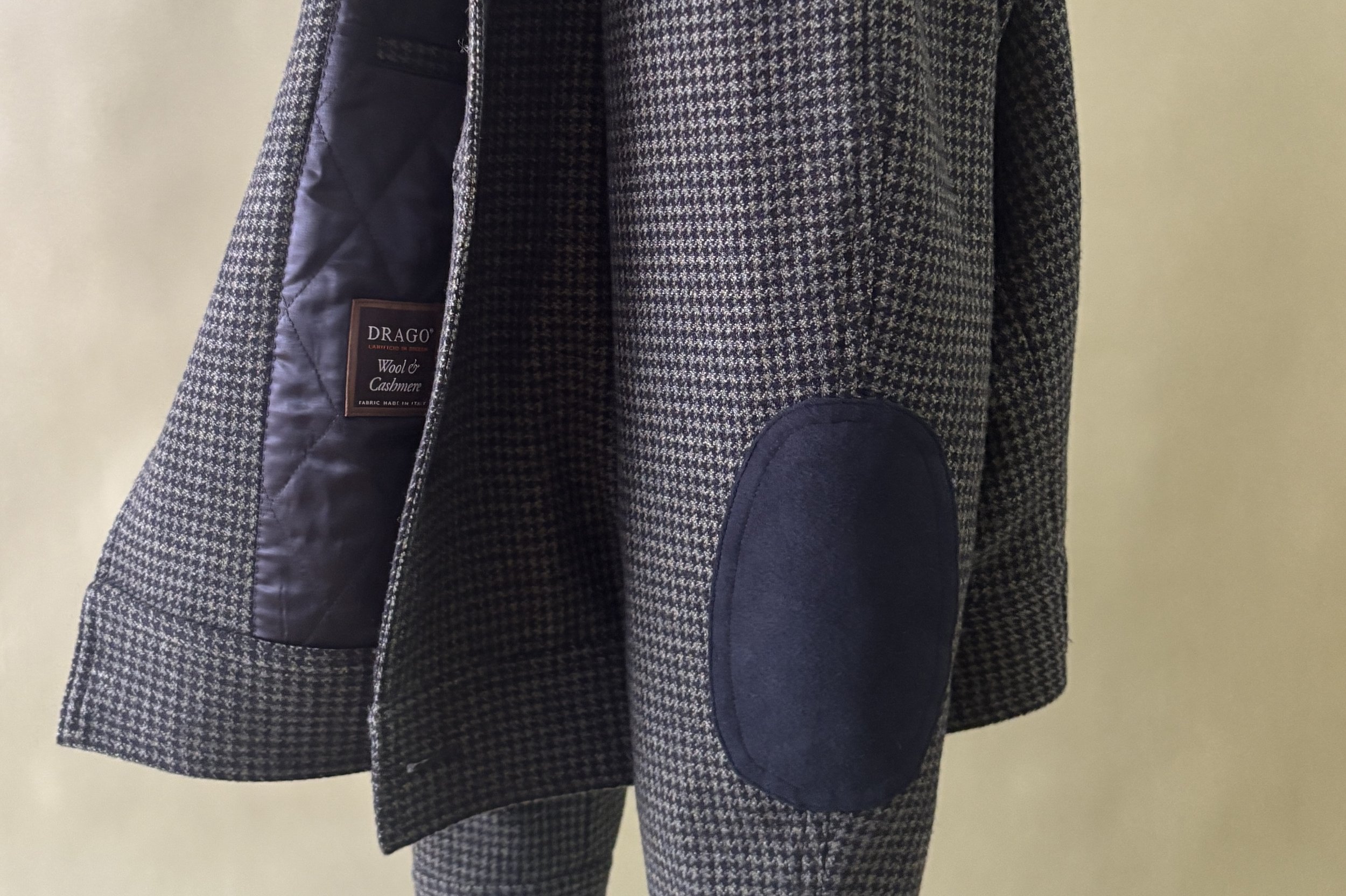 A close-up of a custom made green and black Drago wool & cashmere bomber jacket with a black suede elbow patch against a green backdrop.