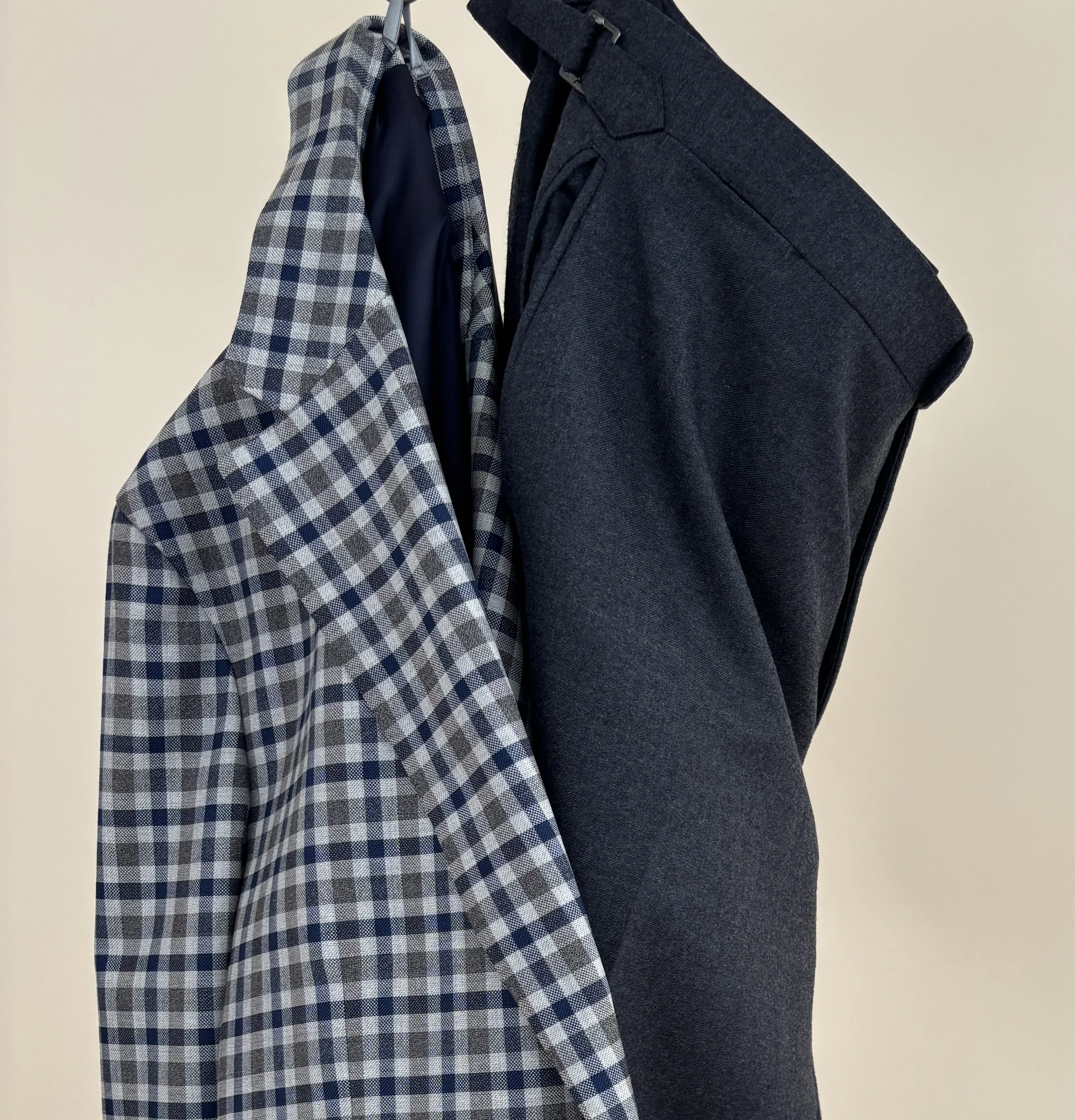 A custom made blue and grey guncheck unconstructed blazer with Vitale Barberis Canonico fabric hanging with custom charcoal grey flannel trousers on a tan background.