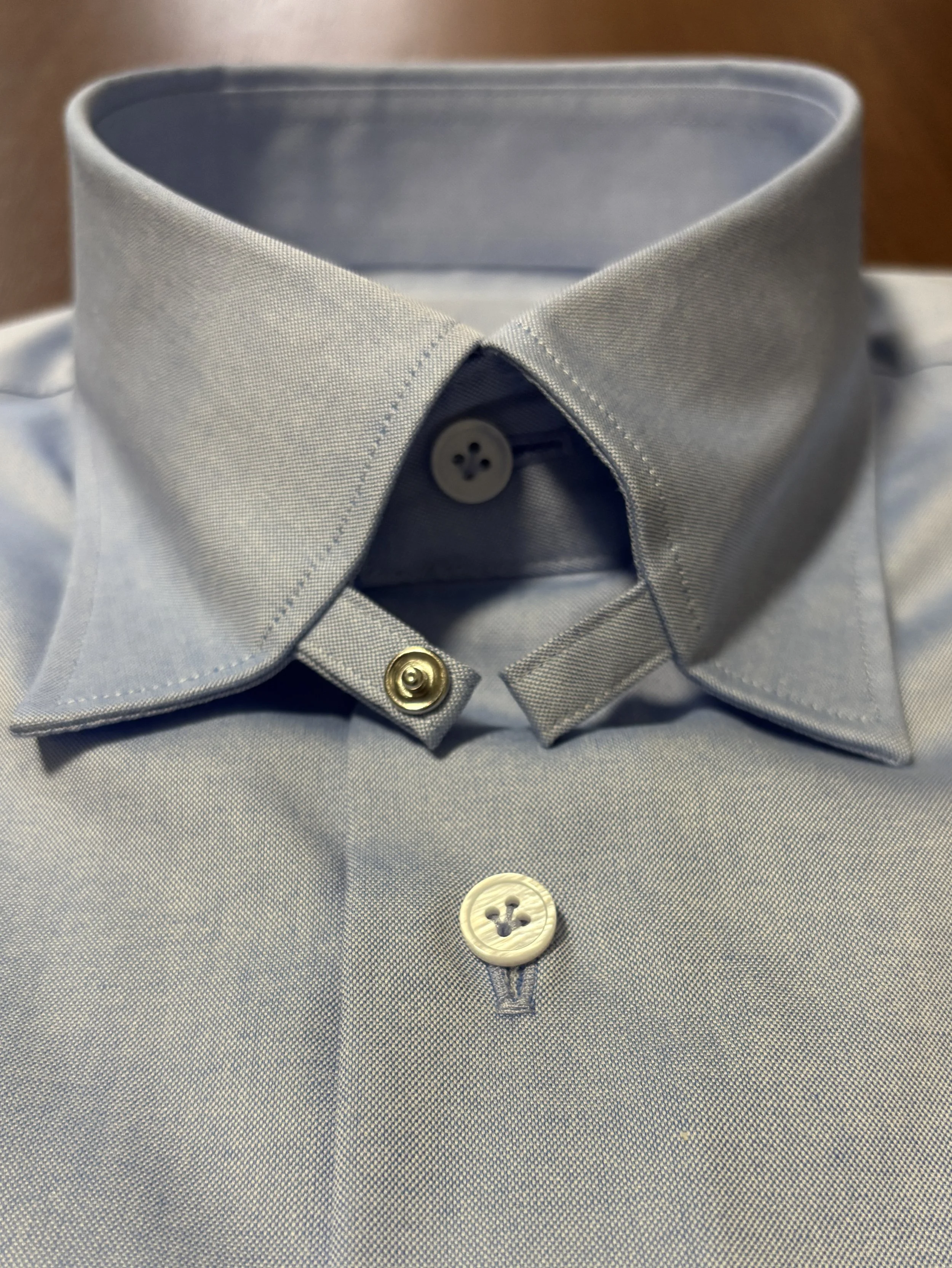 Close-up of a custom made chambray blue dress shirt with a tab collar and mother of pearl shell buttons on a walnut wood table. 