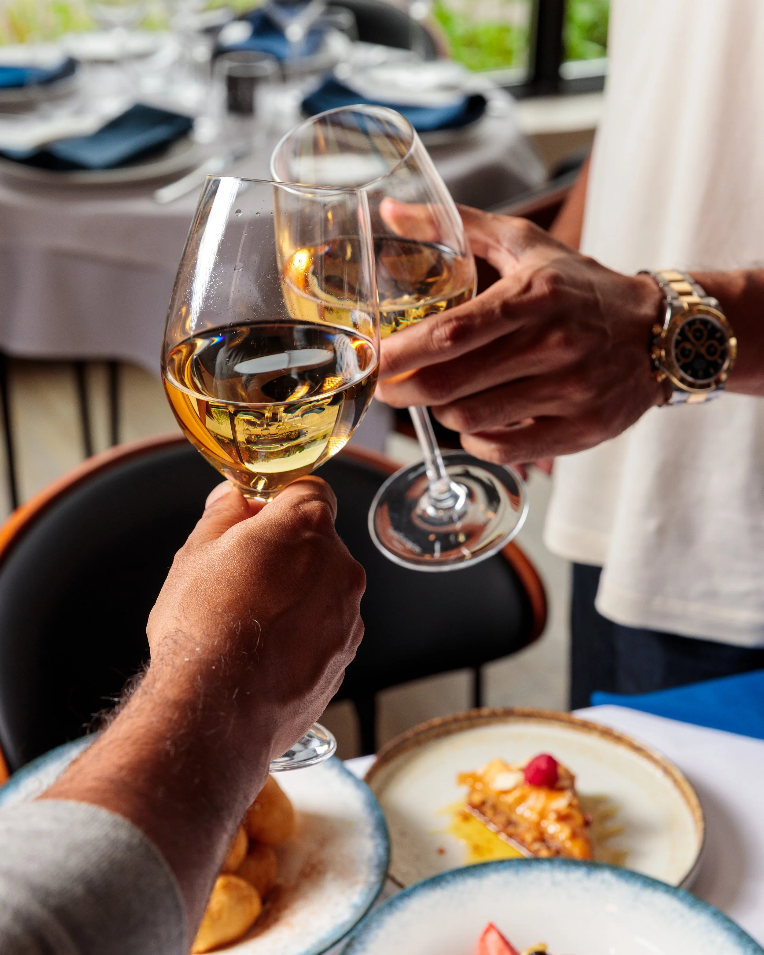 Two people clinking glasses of white wine at a restaurant table with plates of food, including a slice of pie, visible in the foreground.