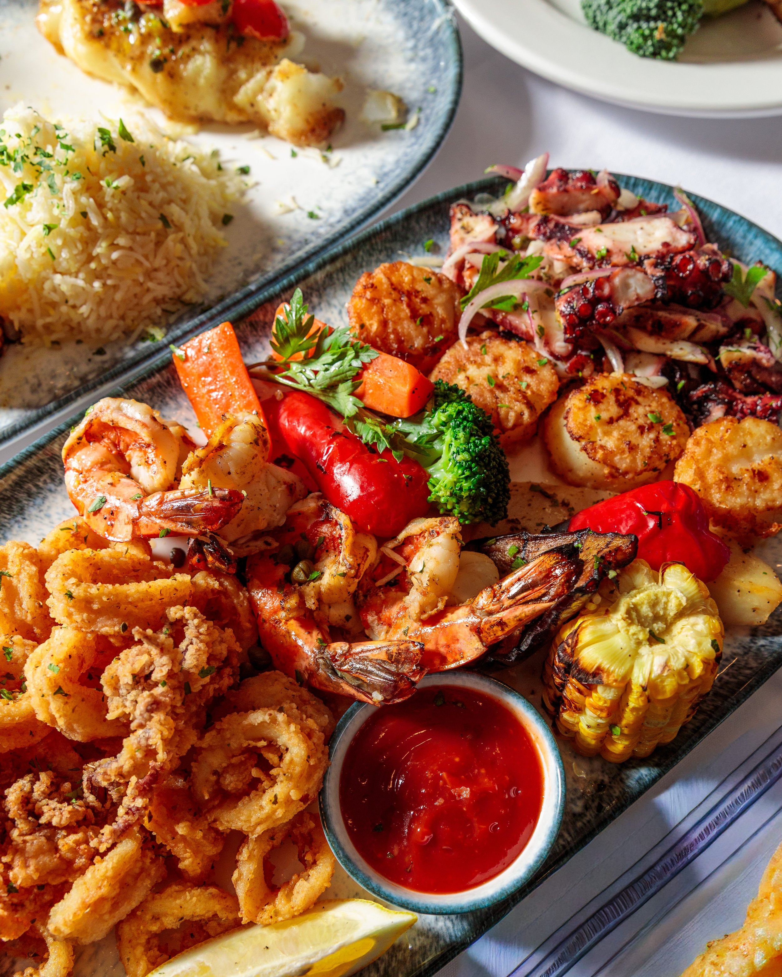 Seafood platter with grilled shrimp, scallops, and calamari, accompanied by grilled corn on the cob, roasted peppers, broccoli, rice, and a side of marinara sauce.