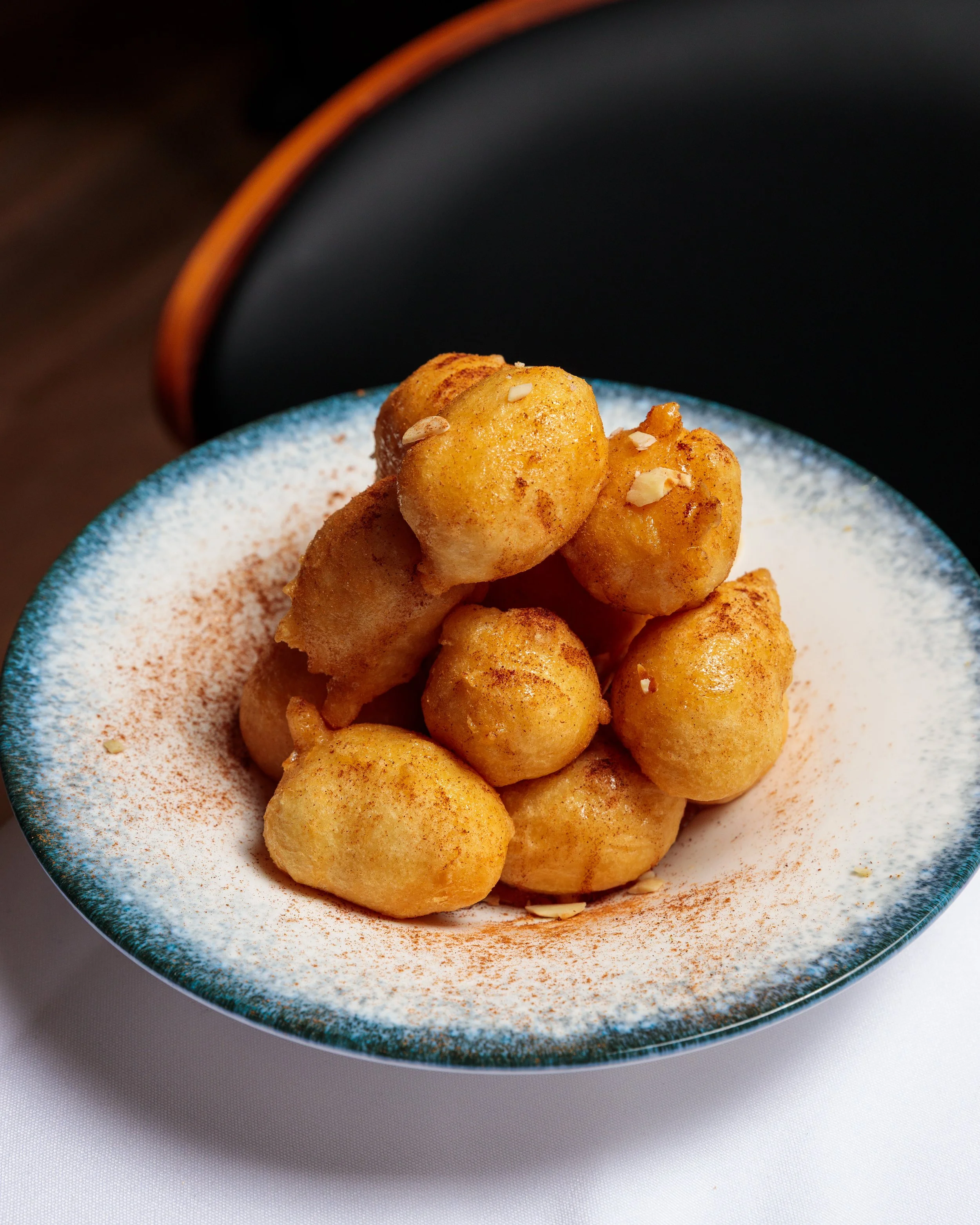 A bowl of fried dough balls topped with chopped nuts and cinnamon, on a white surface.