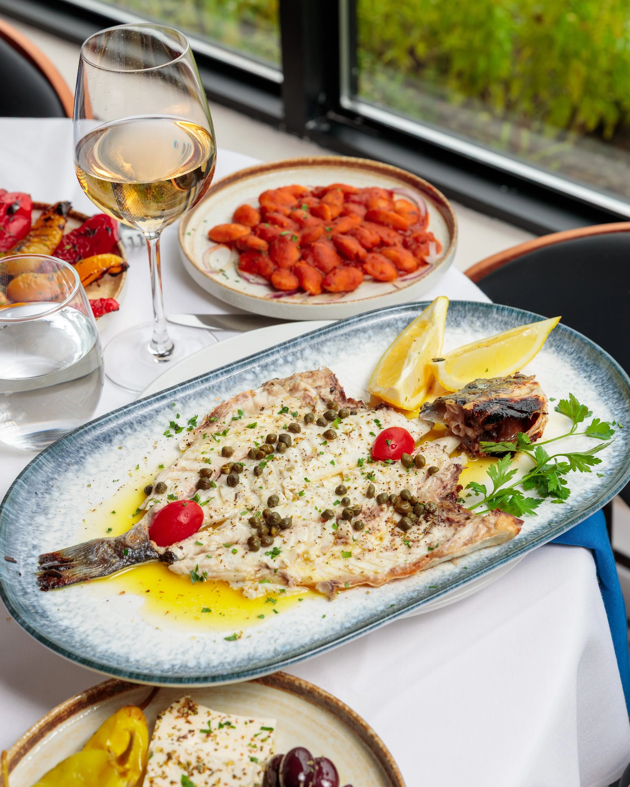 A table with white tablecloth holding a whole grilled fish with lemon wedges, cherry tomatoes, and herbs, a glass of white wine, and plates of various colorful dishes, with a window and greenery outside.