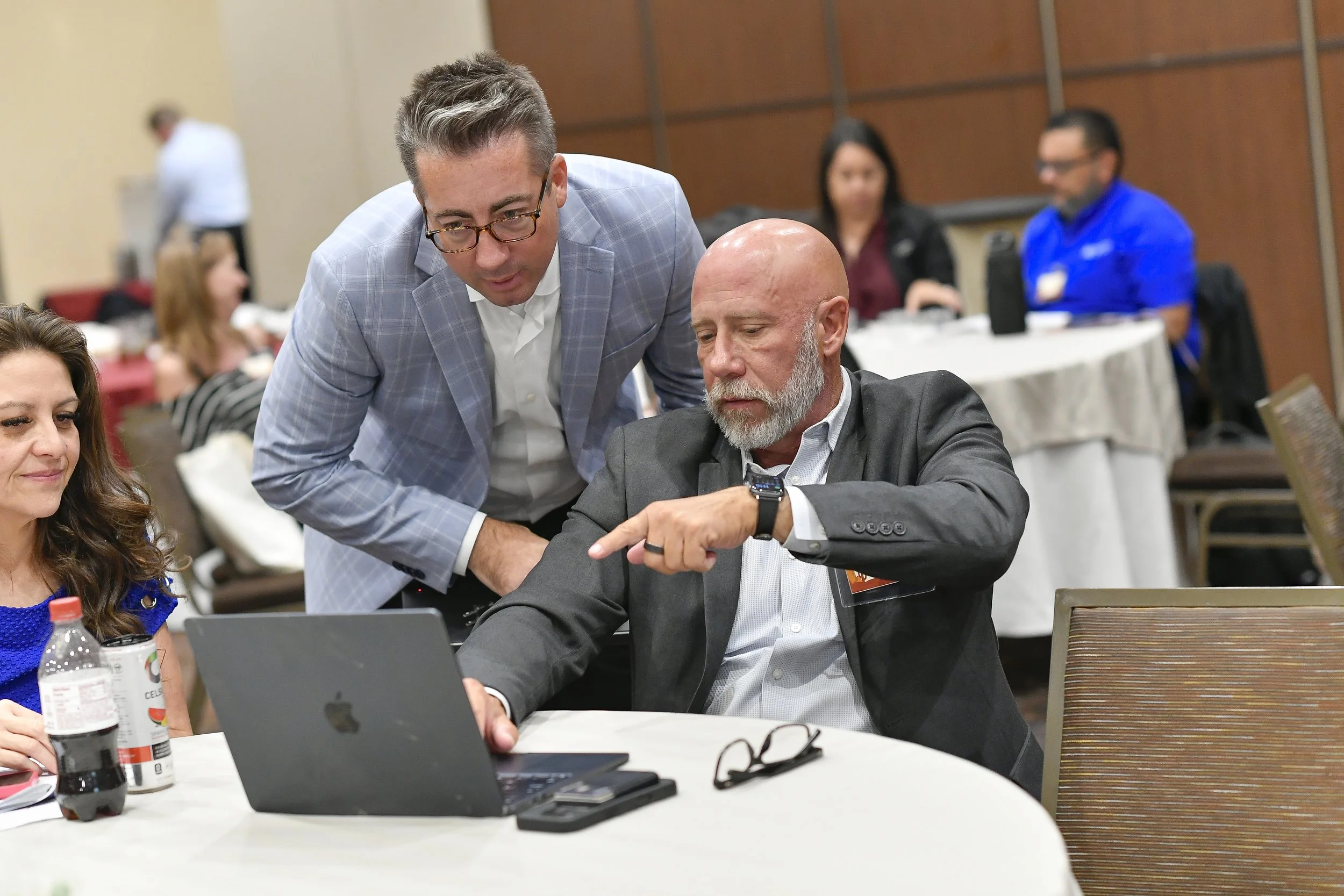 Two men are looking at a laptop screen together during a business event, with one seated and wearing a suit and the other standing and wearing a light gray blazer.