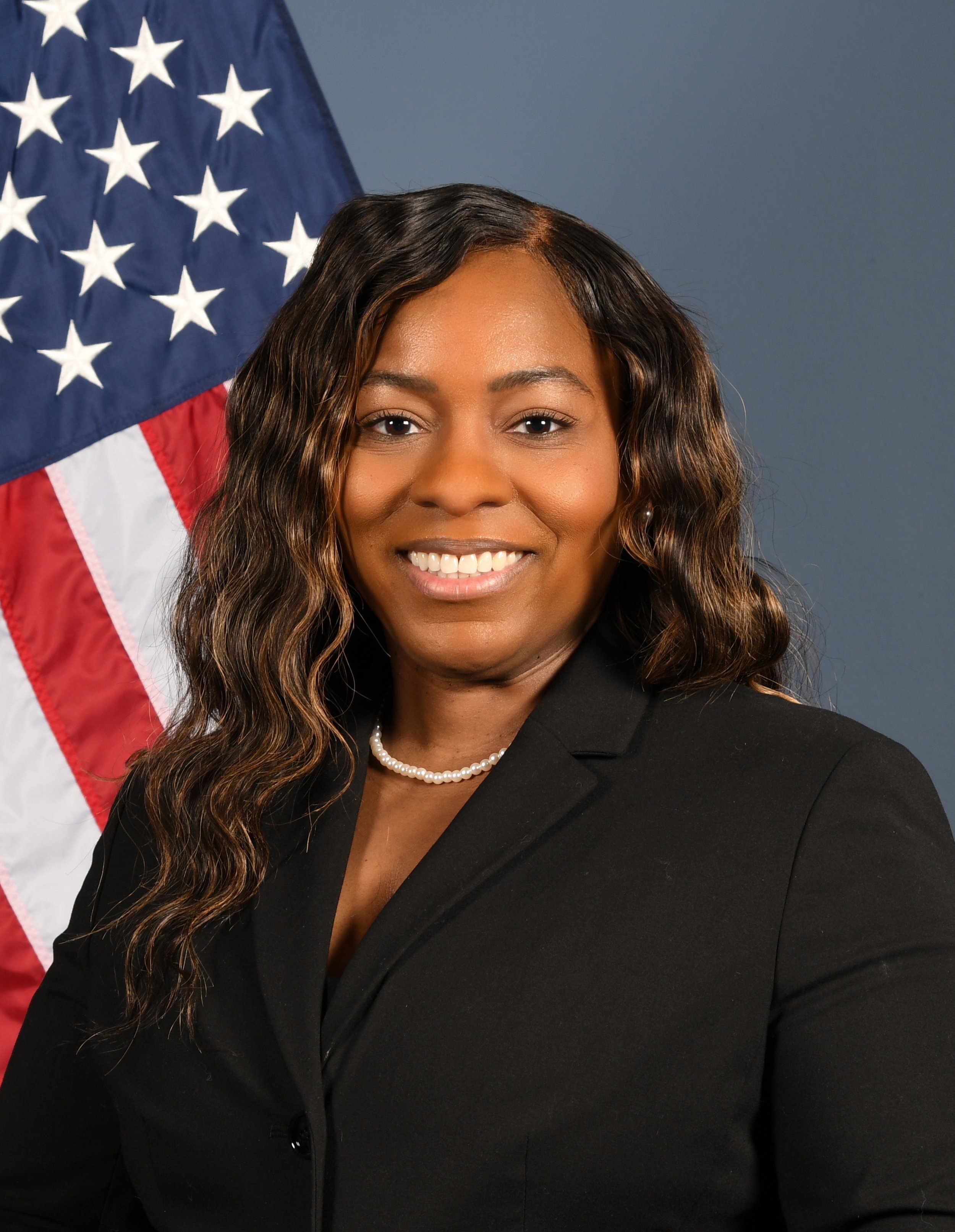 A professional woman with wavy hair and a pearl necklace, dressed in a black blazer, standing in front of an American flag.
