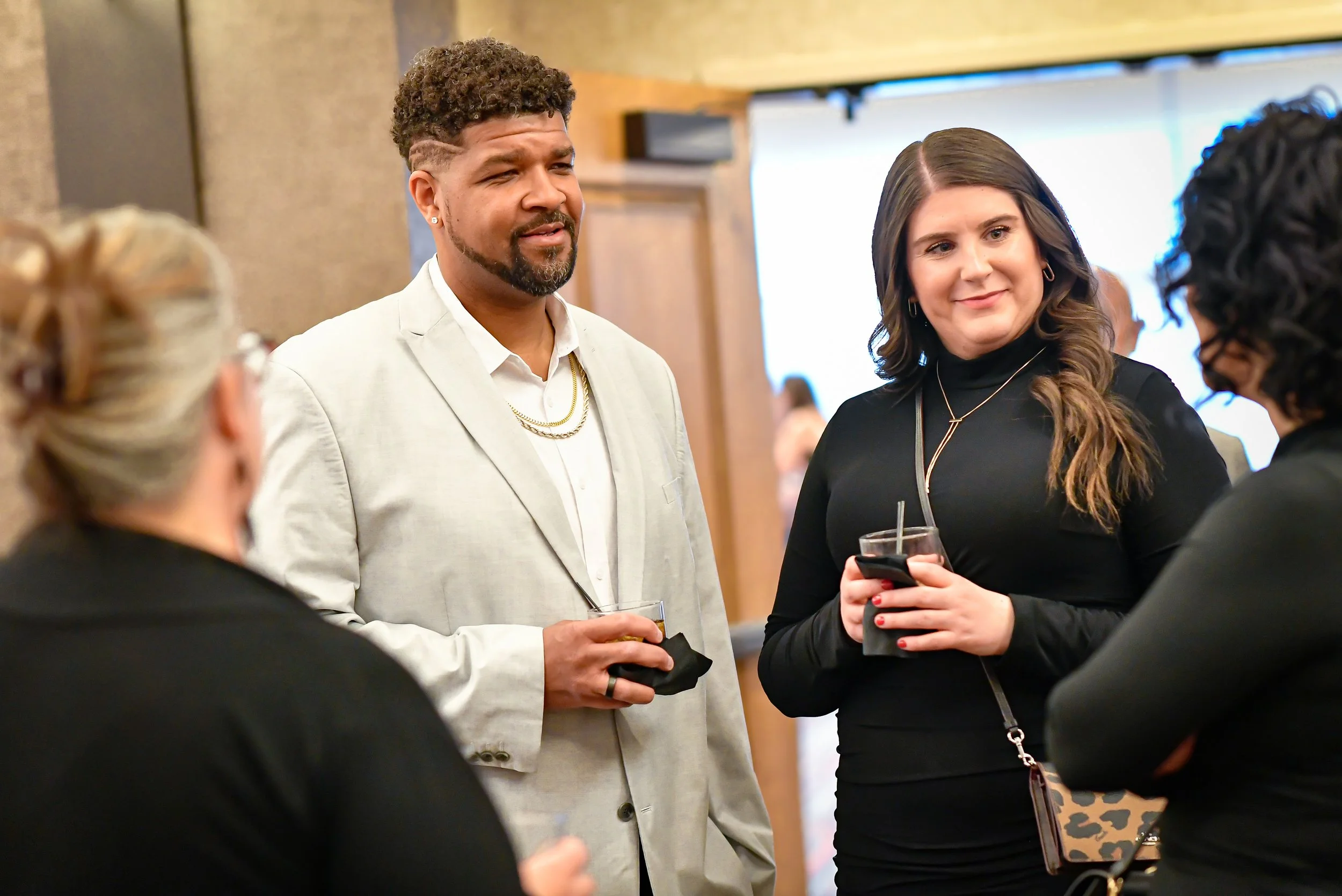 A group of four people having a conversation at a social event indoors. Two women and a man are visible, with another woman partially seen. They are holding drinks, and the woman on the right has a phone in hand. The background is blurred with warm lighting.