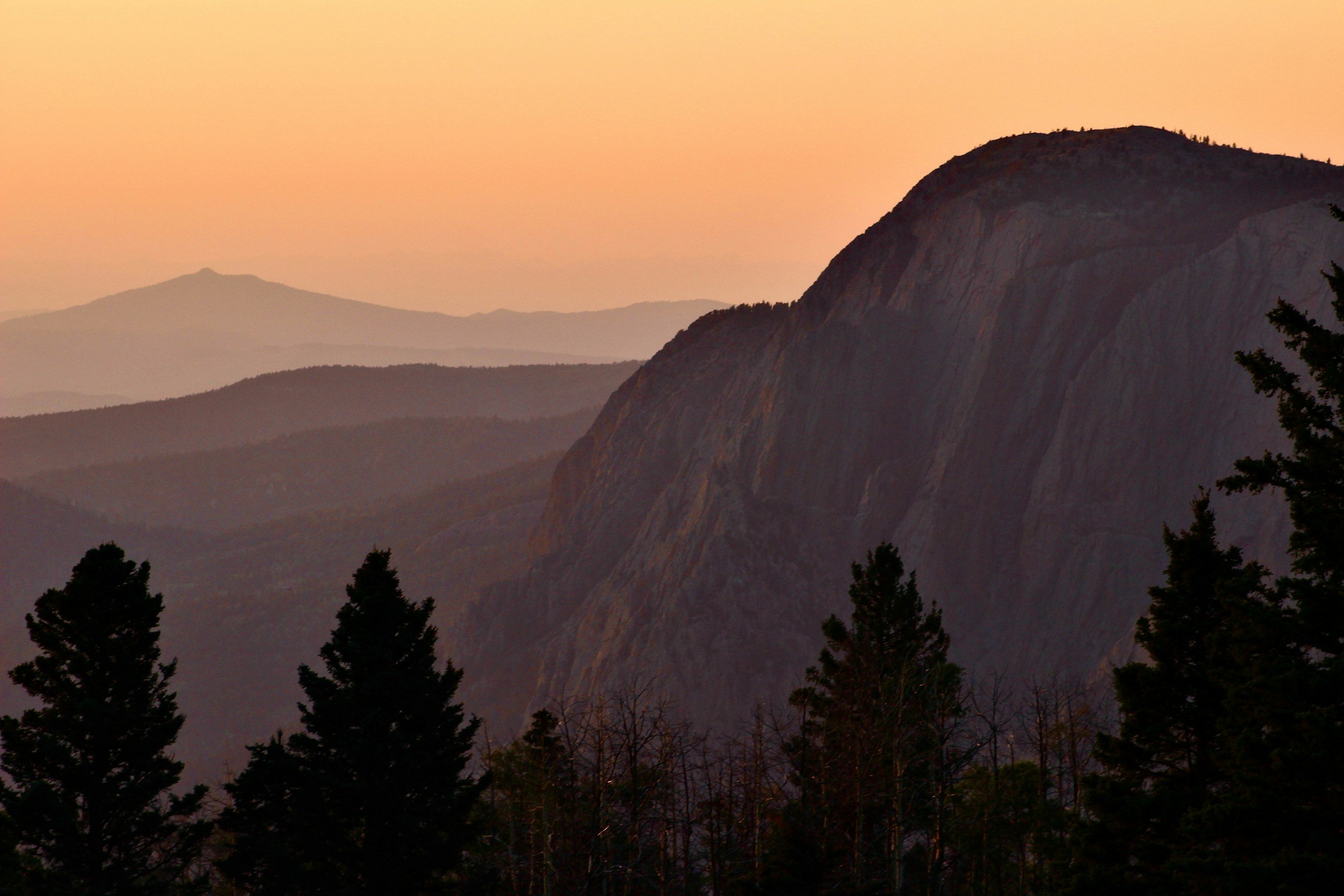 Mountain landscape during sunset with layers of mountains and trees in the foreground.