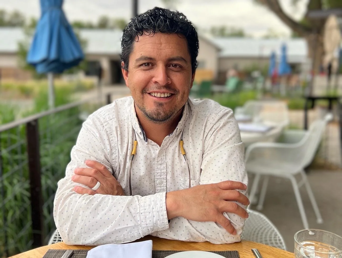 A man with dark curly hair, wearing a white shirt with small dots, smiling and sitting at an outdoor restaurant table. There is a glass of water and a napkin on the table, with green plants, other tables, and umbrellas in the background.