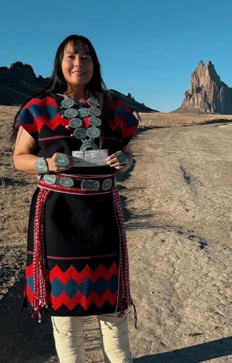 A woman dressed in traditional Native American attire with jewelry, standing outdoors in a desert landscape with a mountain in the background.