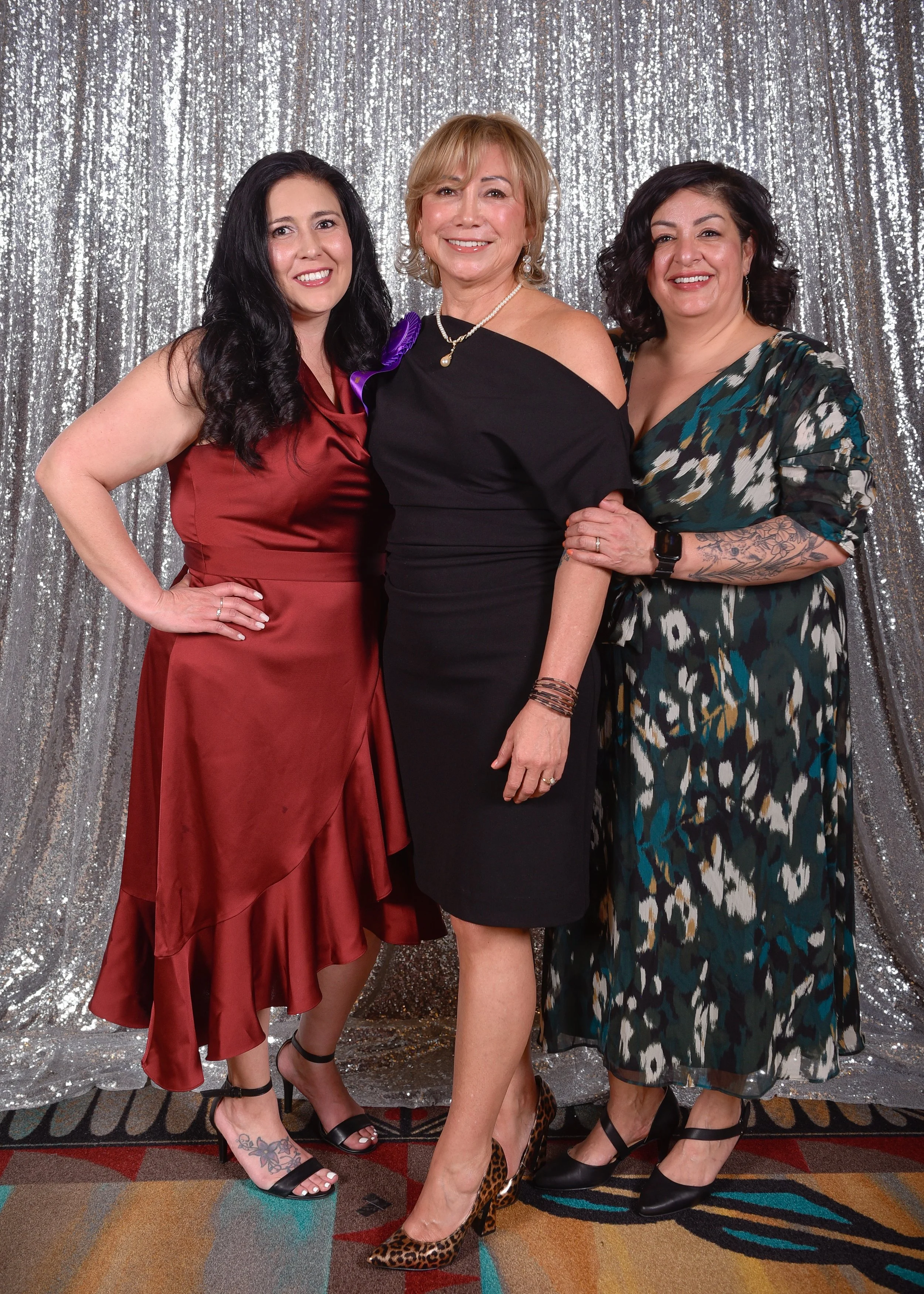Three women standing together in front of a silver sequin backdrop, smiling at the camera. All wearing elegant dresses and heels, with their arms around each other.