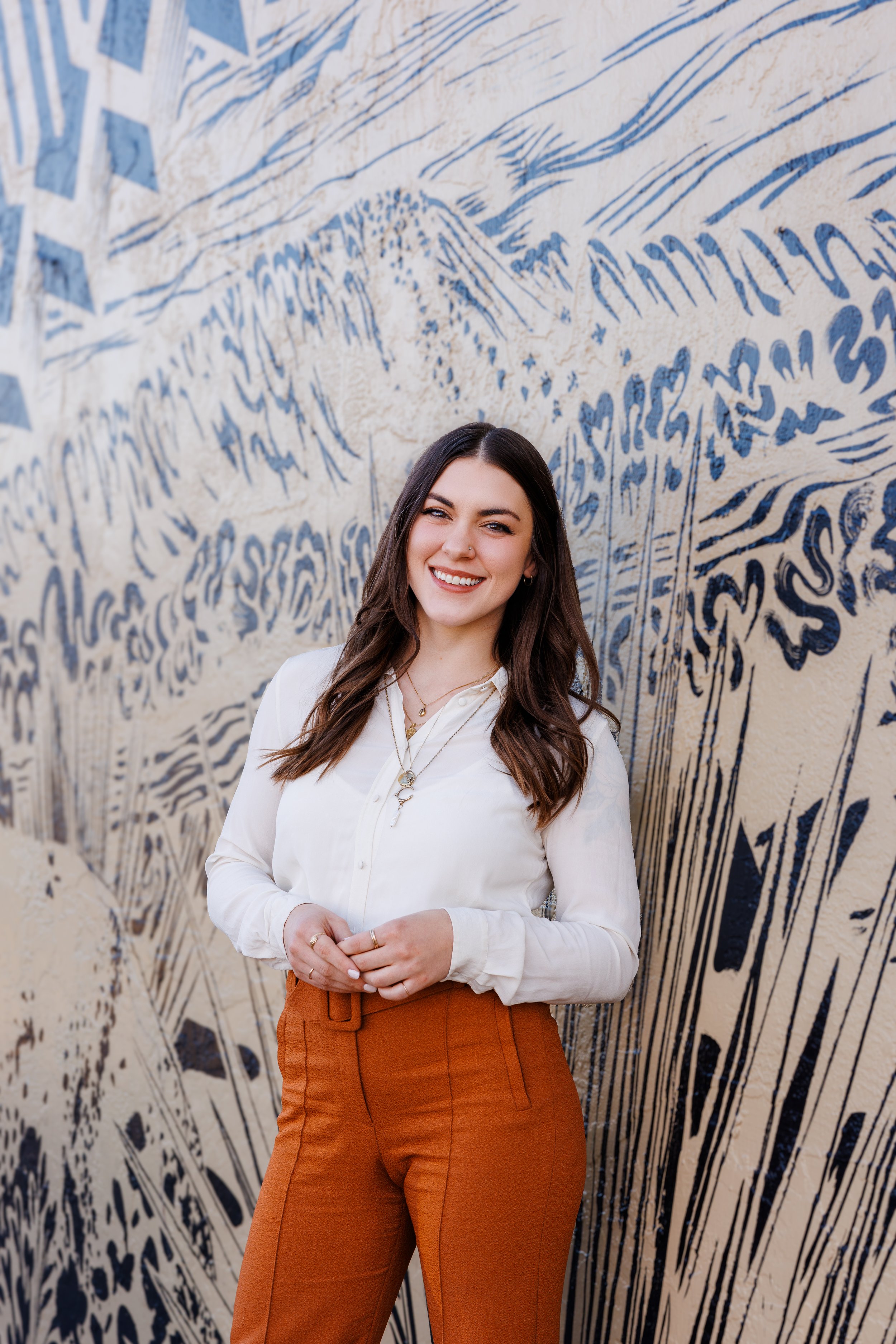 Young woman smiling, wearing a white blouse and orange pants, standing in front of a beige wall with blue abstract mural art.
