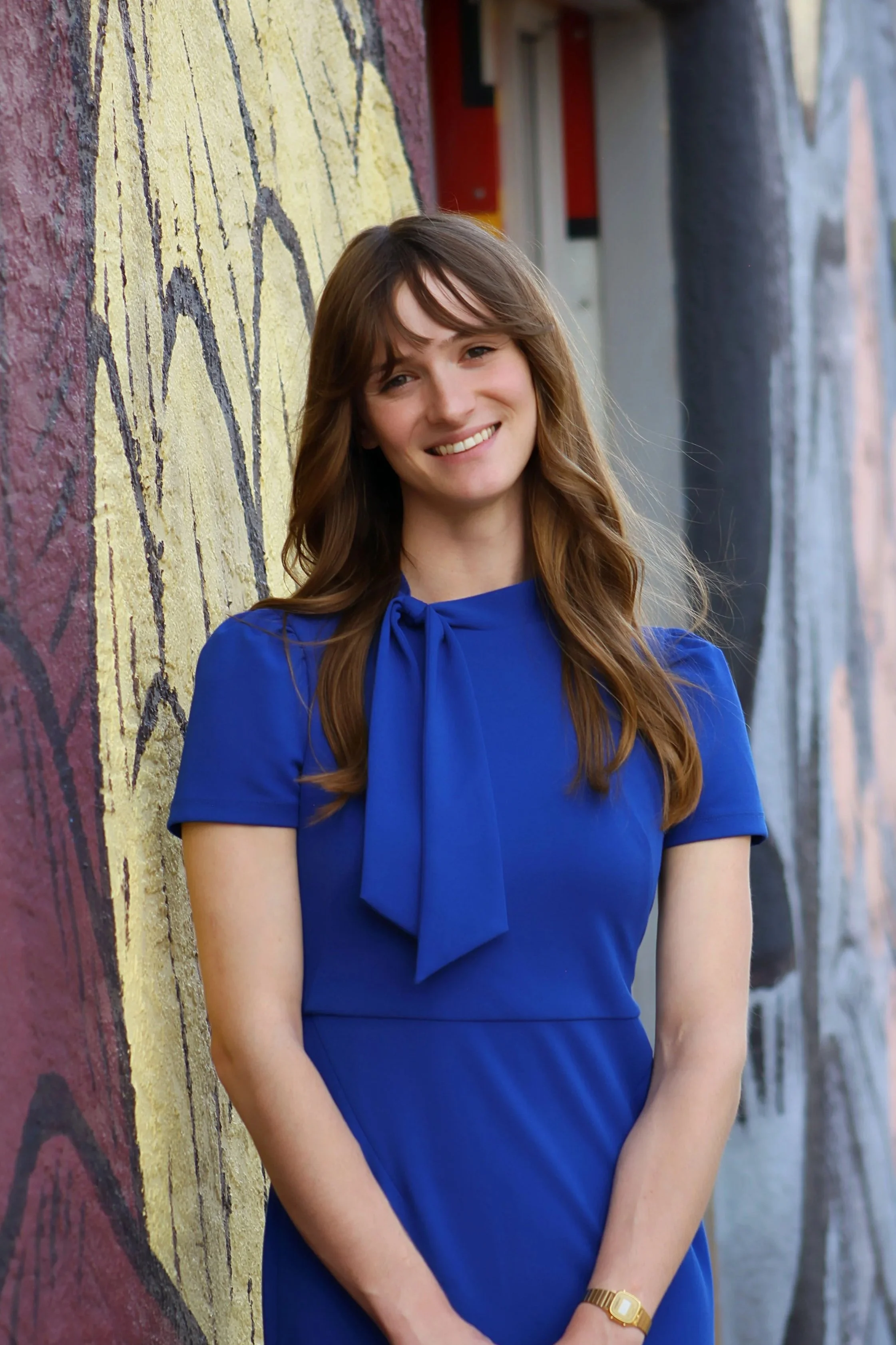 A young woman with long wavy brown hair, wearing a light blue button-up shirt, smiling outdoors with green foliage and white flowers in the background.