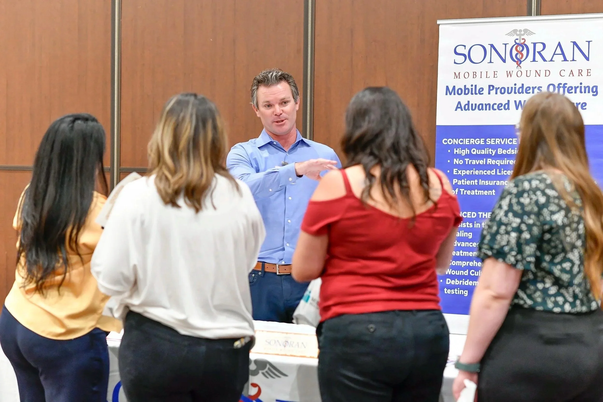 Man explaining products to a group of women at a marketing booth in front of a Sonoran Wound Care banner.