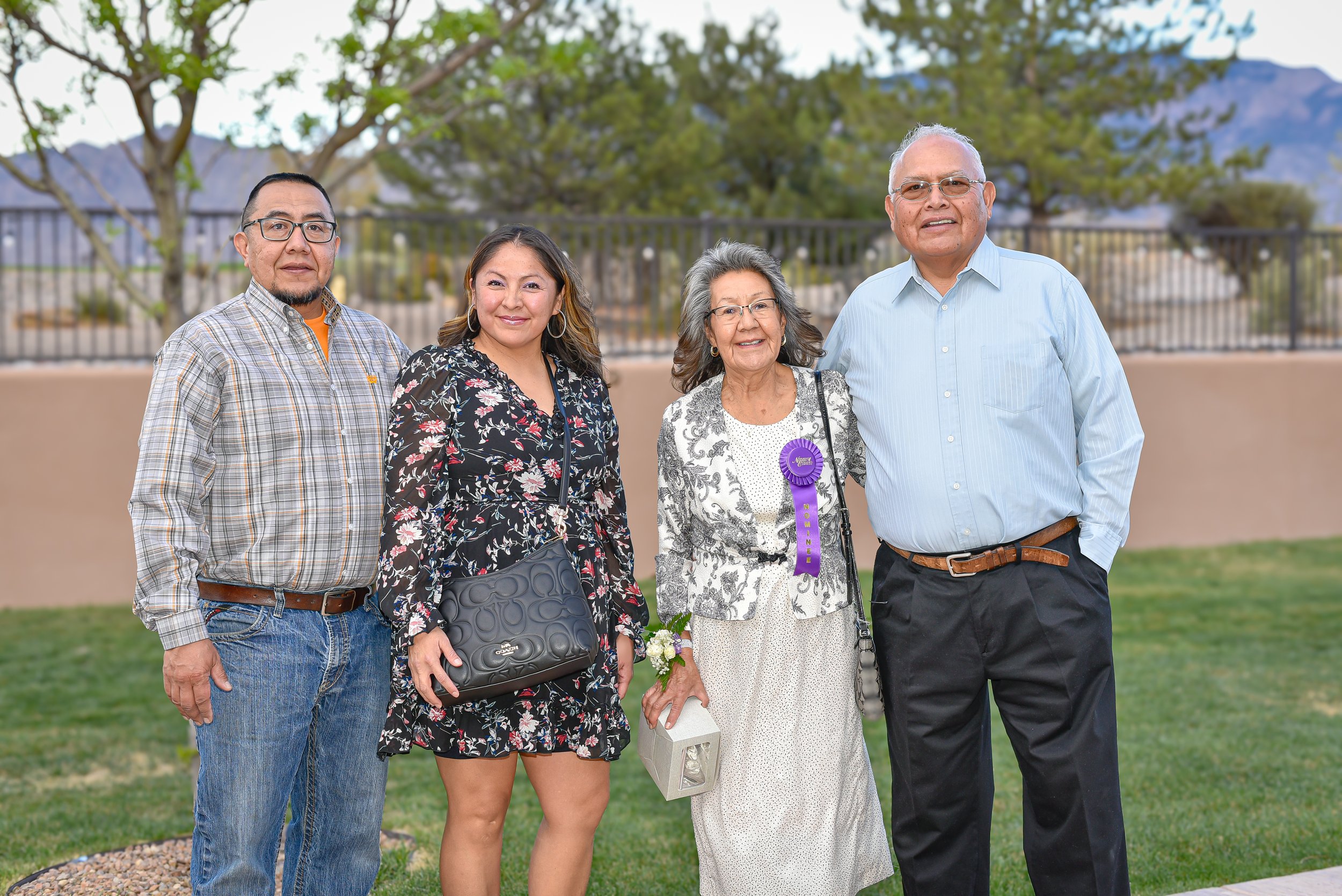 Group of five people standing outdoors, four adults and one elderly woman with a purple ribbon, in front of a fence with trees and mountains in the background.