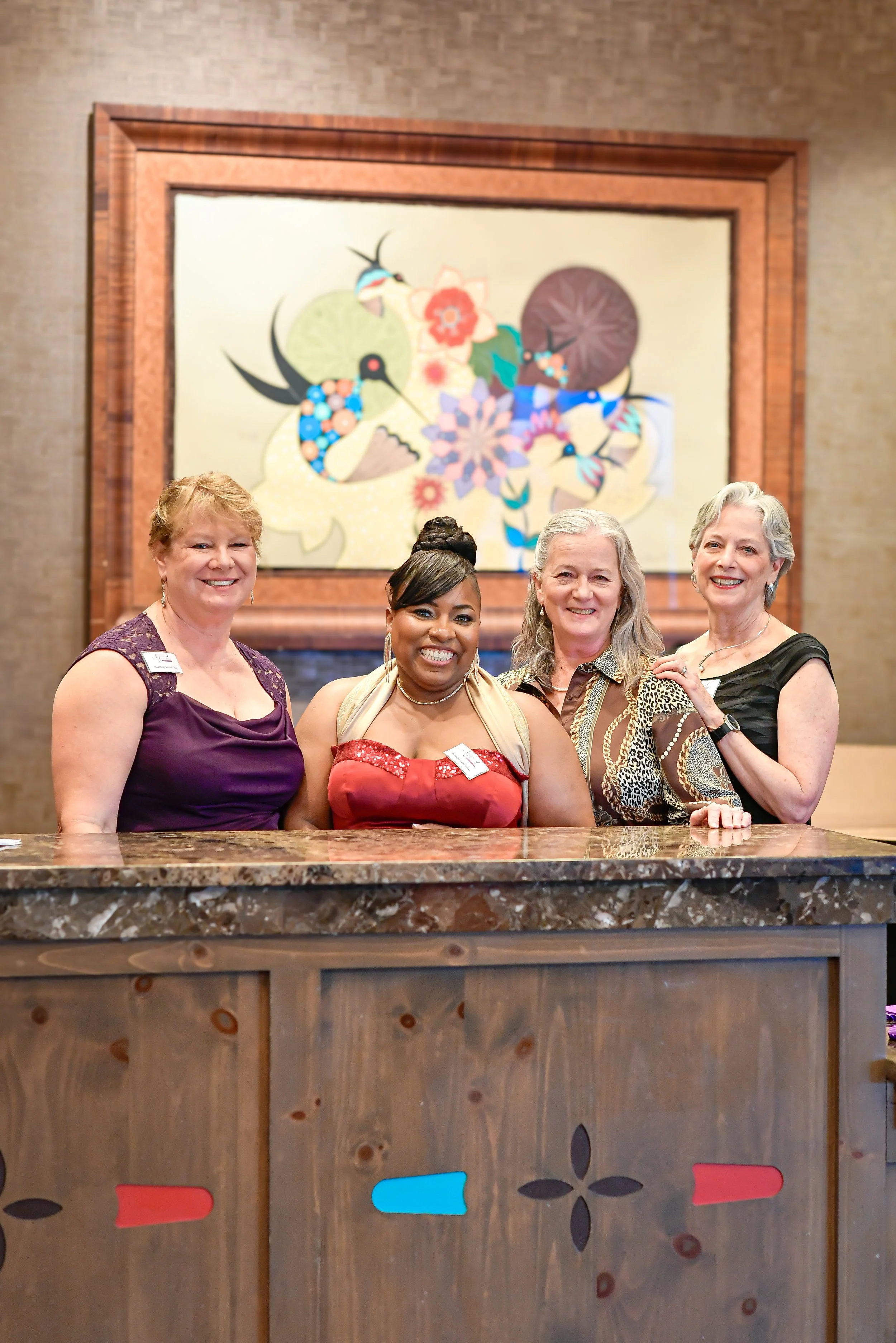 Four women standing behind a marble reception desk, smiling at the camera, with an abstract artwork featuring birds and flowers hanging on the wall behind them.