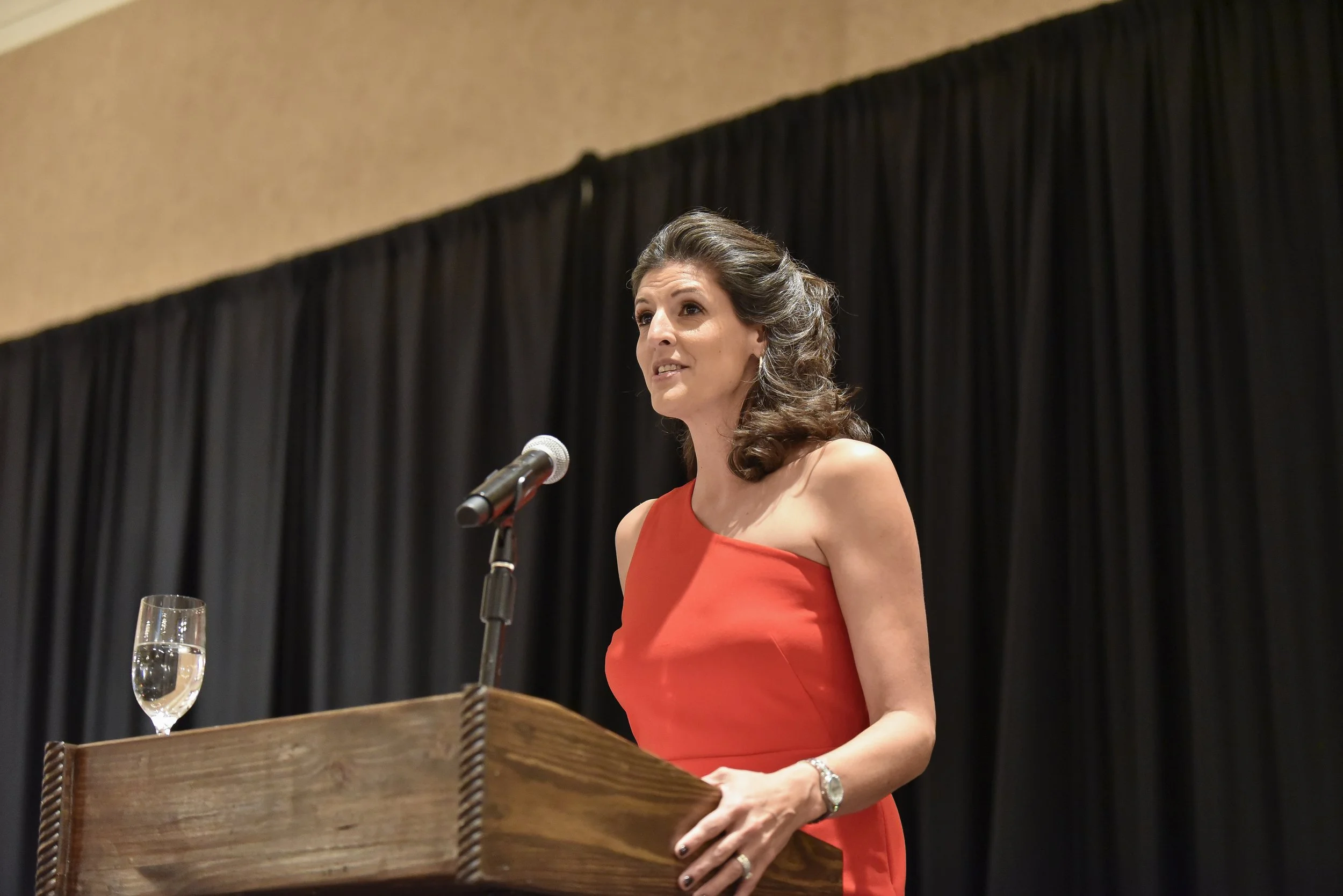 A woman in a red dress speaking at a podium with a microphone, a glass of water, and a black curtain background.