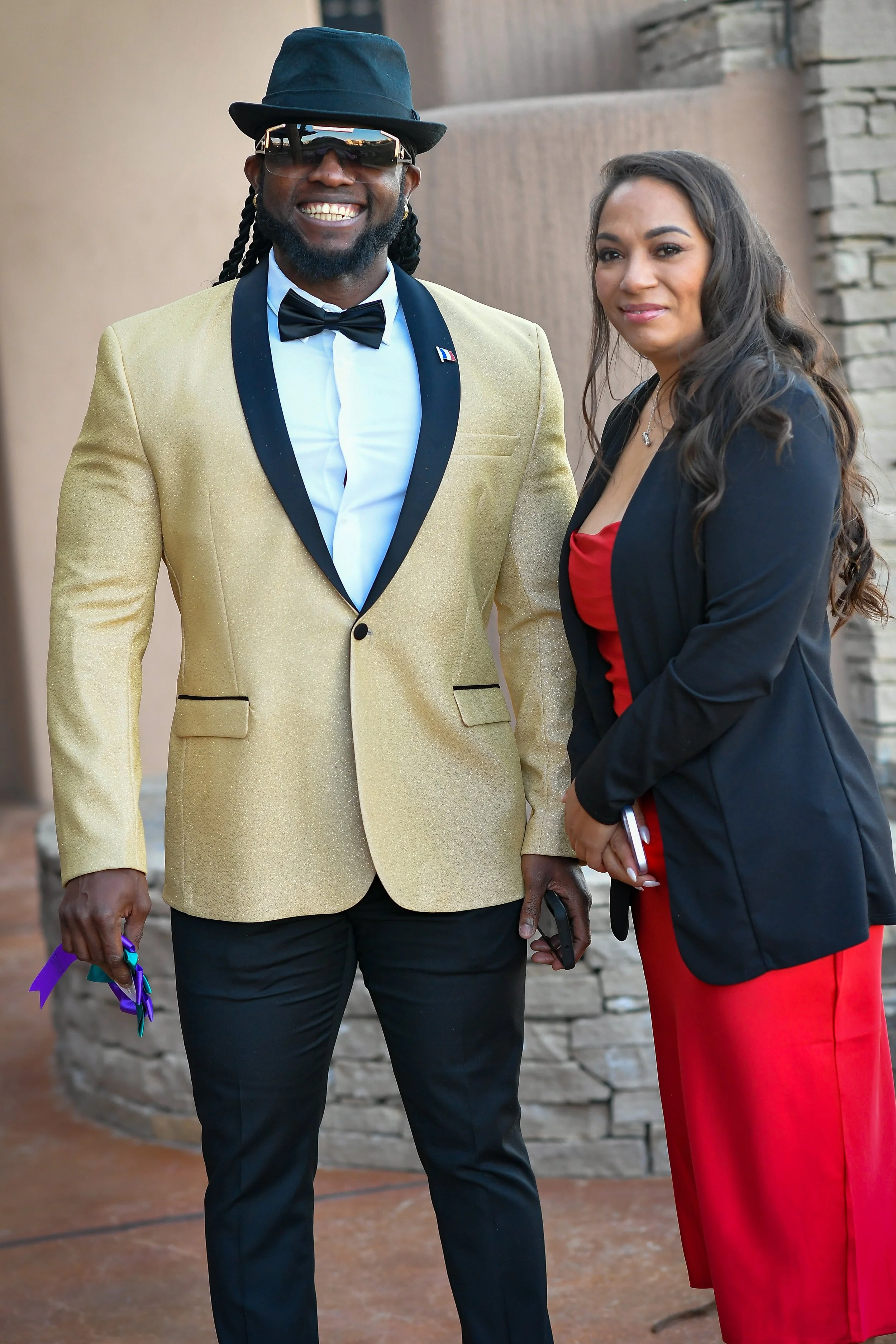 A man in a gold tuxedo jacket, black bow tie, black pants, wearing sunglasses and a hat, standing beside a woman in a black blazer and red dress, outdoors near a stone wall.
