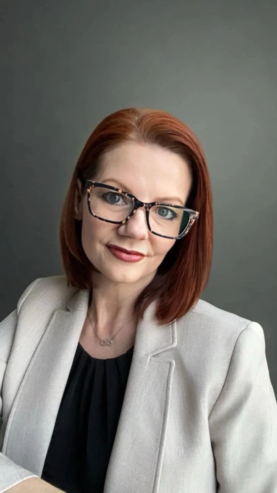A woman with shoulder-length red hair wearing eyeglasses, a white blazer, and a black top, smiling at the camera against a plain gray background.