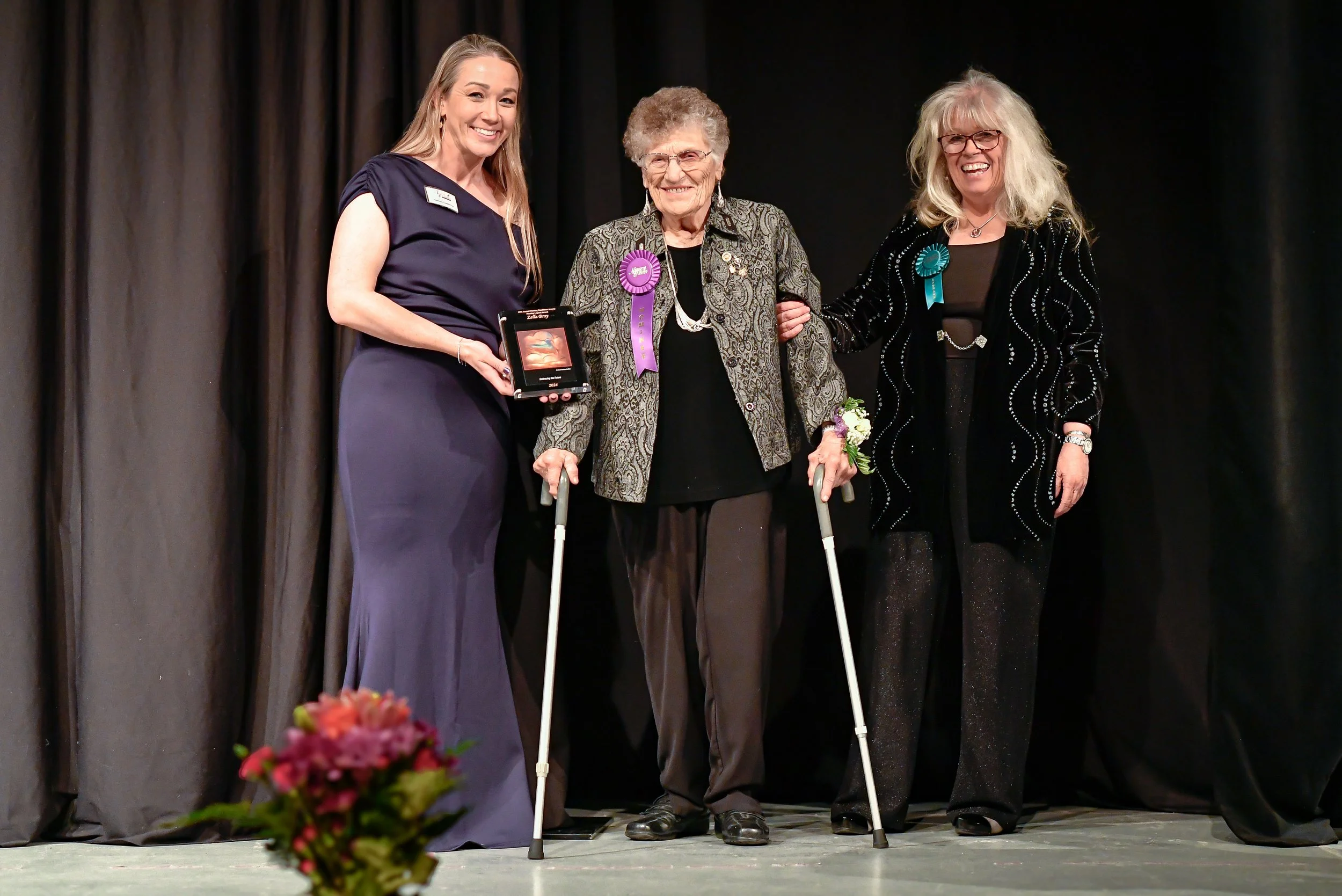 Three women on stage, with one elderly woman in the middle holding crutches, being awarded or recognized at an event. The elderly woman has purple ribbon badges on her jacket, and the other two women are smiling and dressed in formal attire.